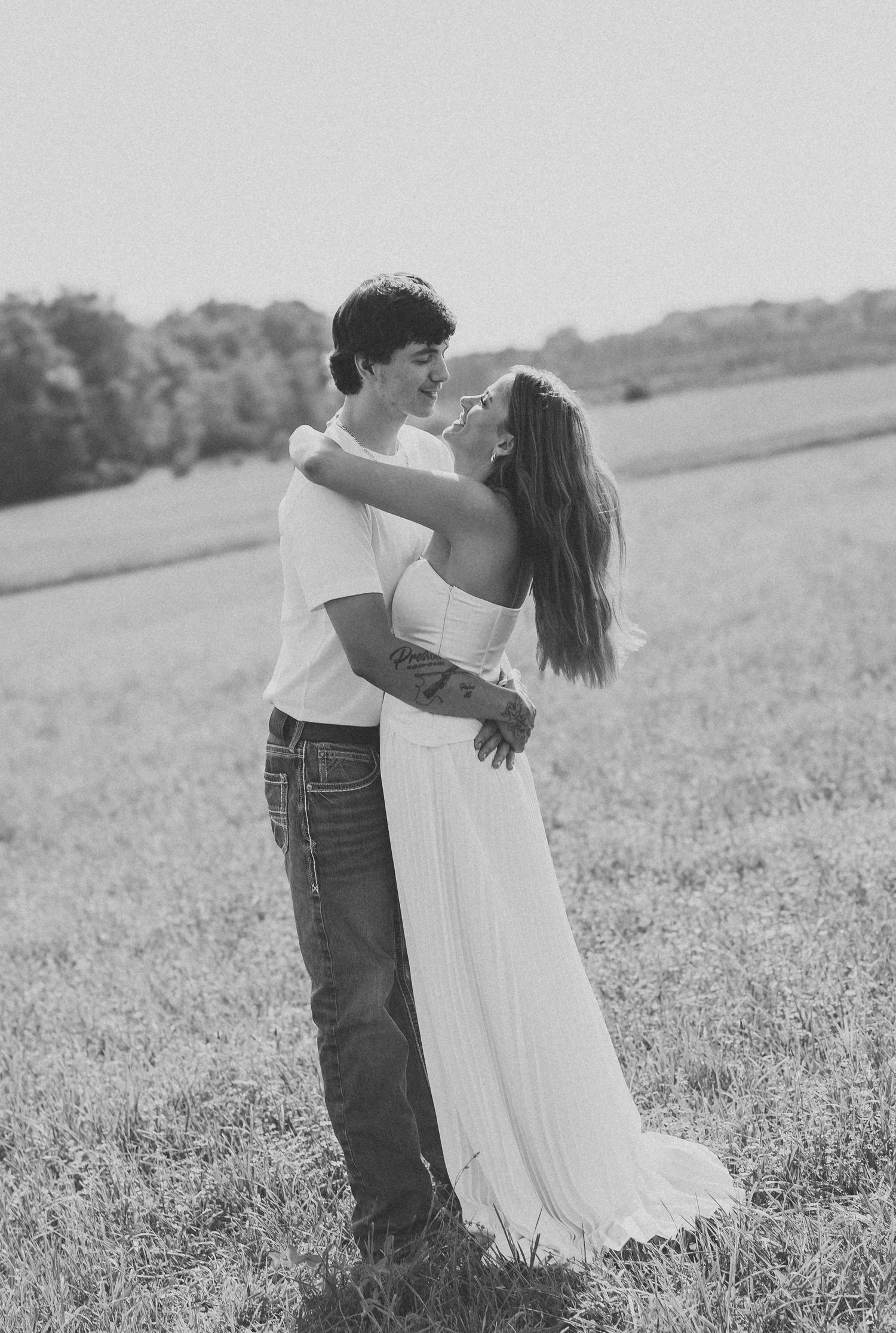 Black and white photo of a young couple embracing in a field, woman in white dress gazing up at man.