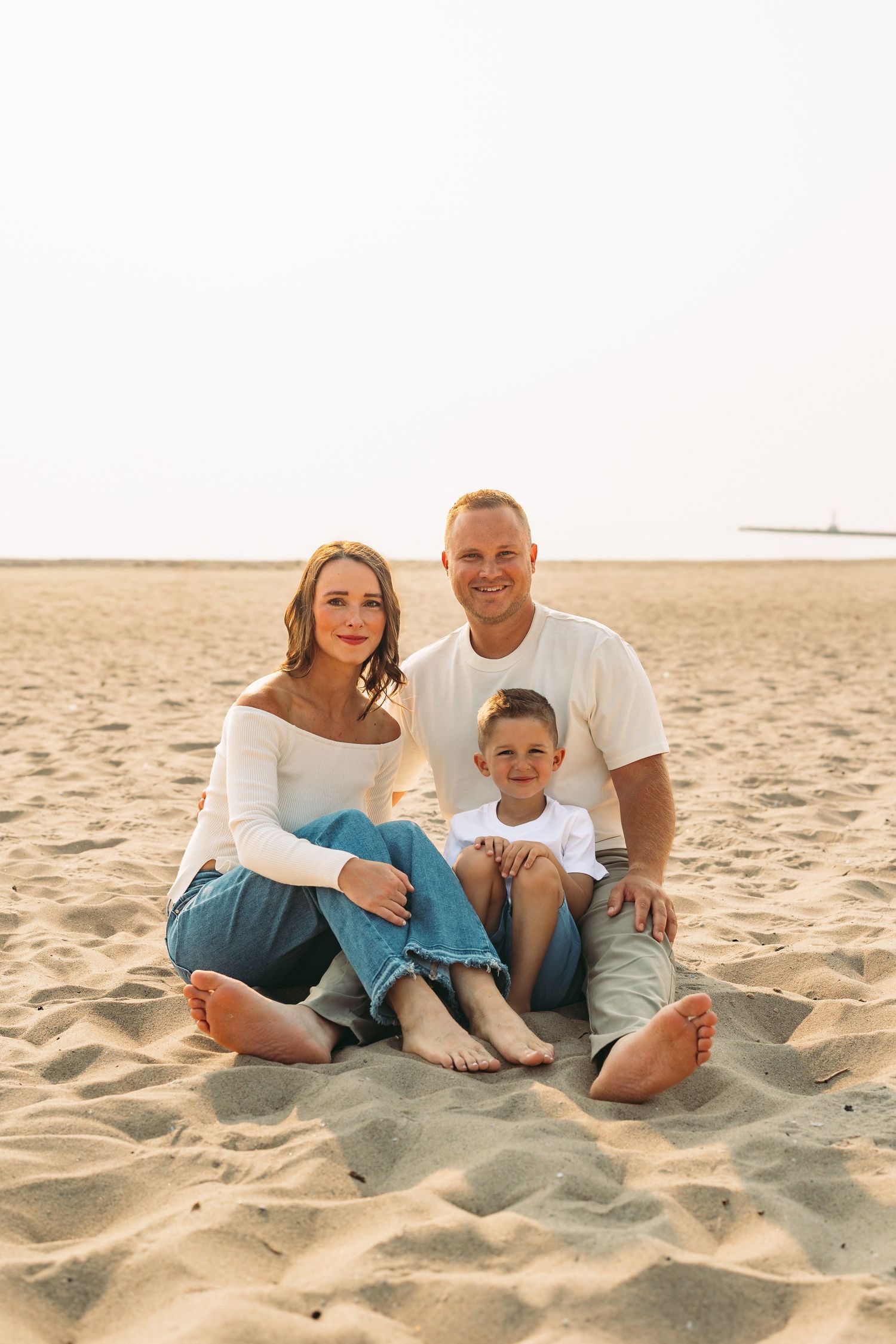 Happy family of three sitting barefoot on sandy beach, smiling couple with young child in casual white tops and jeans.