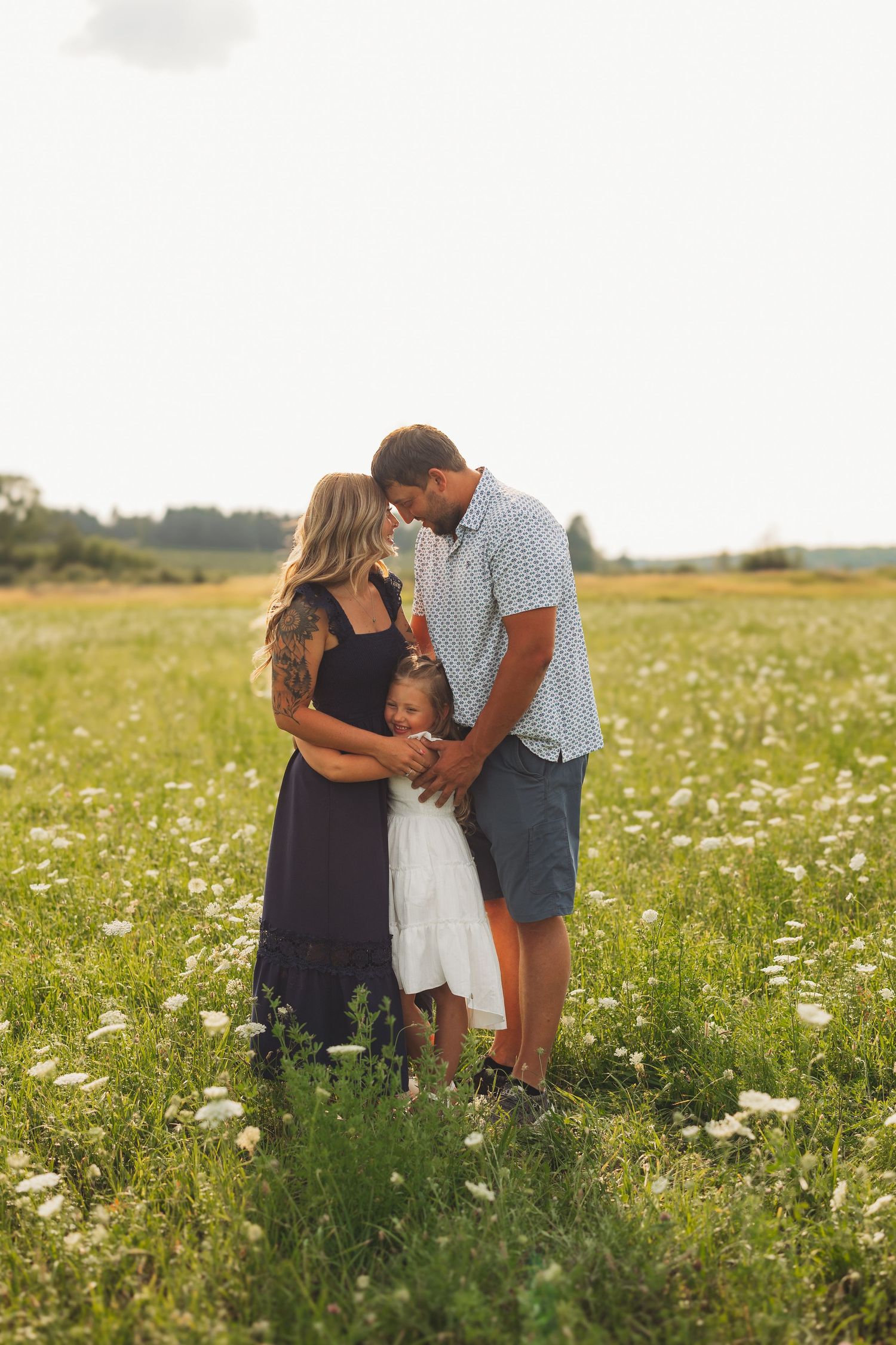 Family of three standing in a sunny wildflower field, parents embracing young daughter in white dress.
