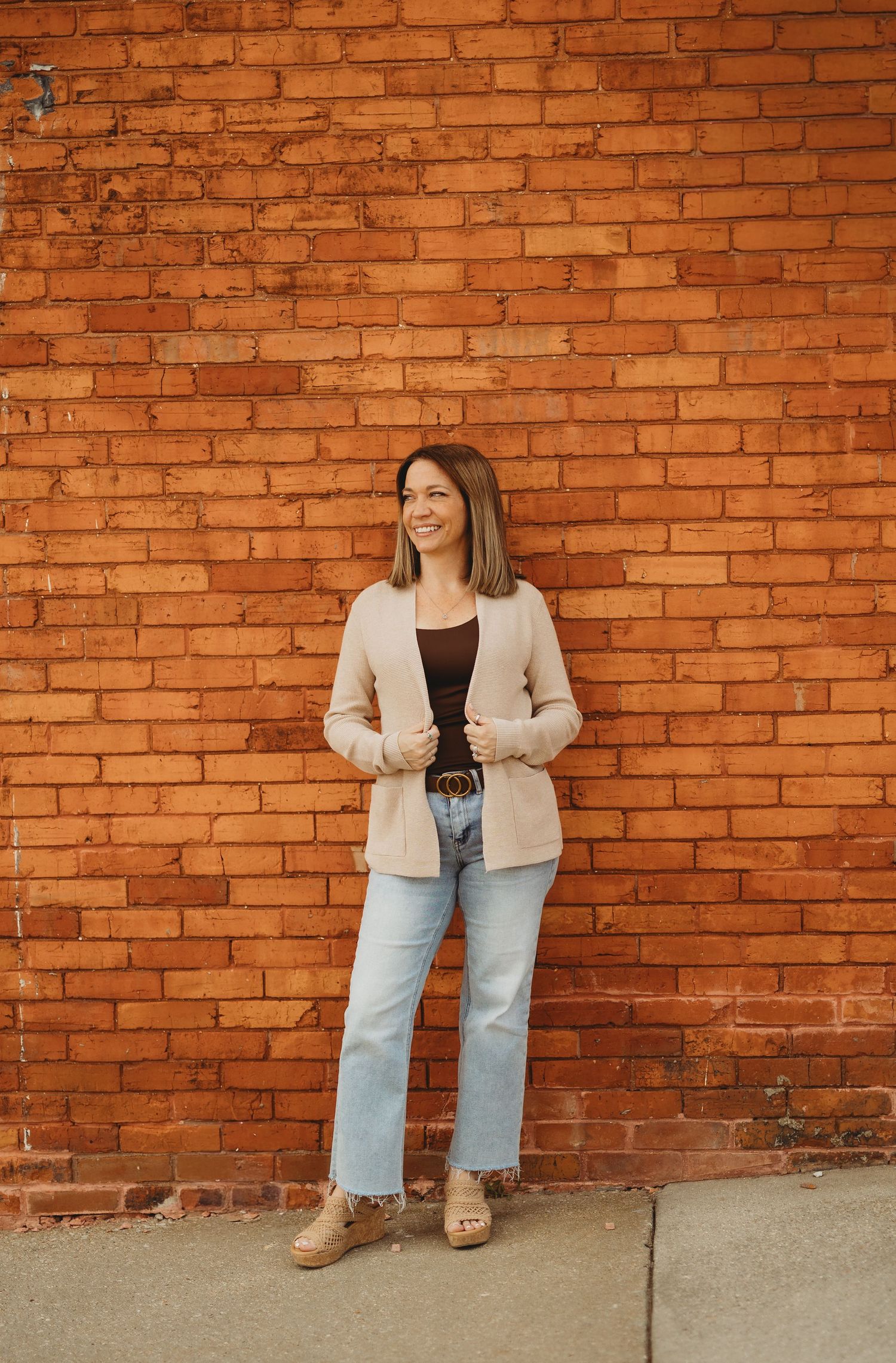 Woman in beige blazer, dark top, and light jeans smiling against a rustic red brick wall.