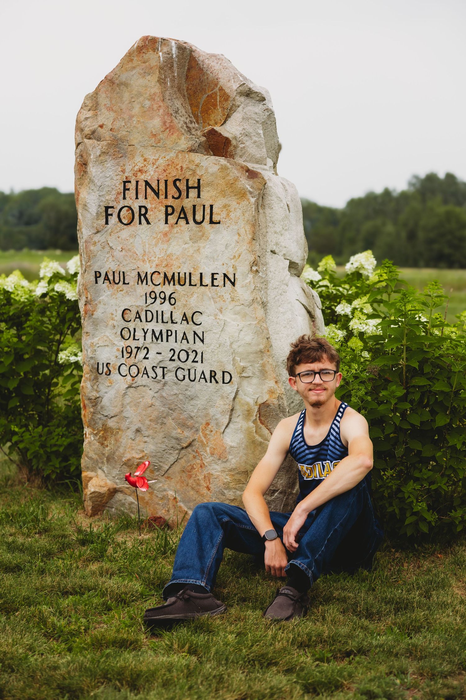 Young man in a striped running singlet sits beside a stone memorial reading Finish for Paul, honoring Olympian Paul McMullen.
