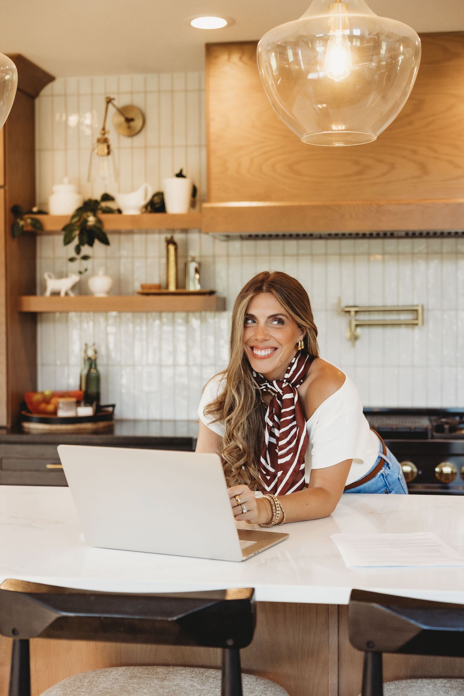 Smiling woman in a striped scarf leans on a white kitchen island while working on a laptop under glass pendant lights.