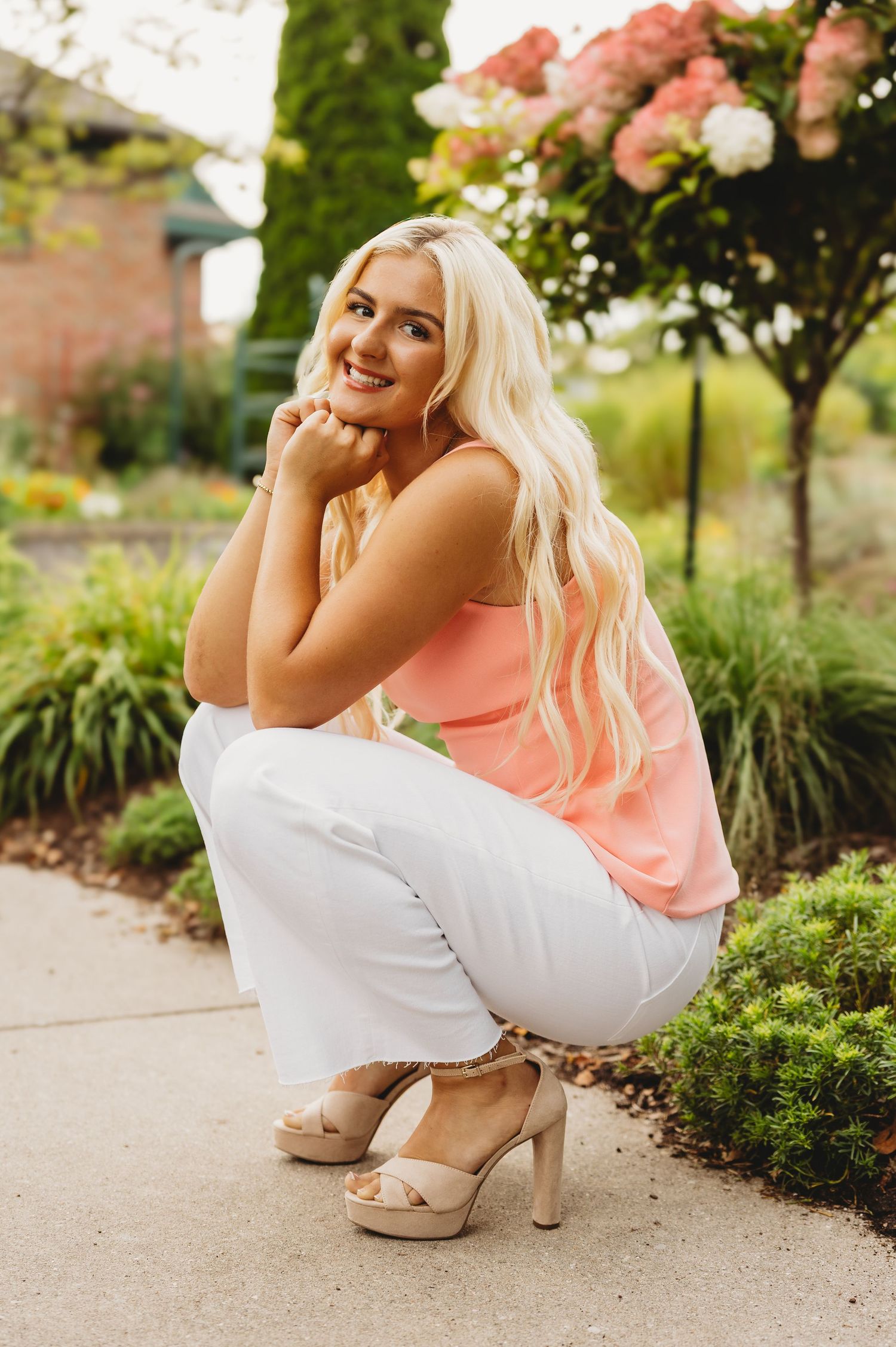 Smiling woman with long blonde hair crouches on a garden path in a peach top, white pants and nude heels near blooming pink flowers.
