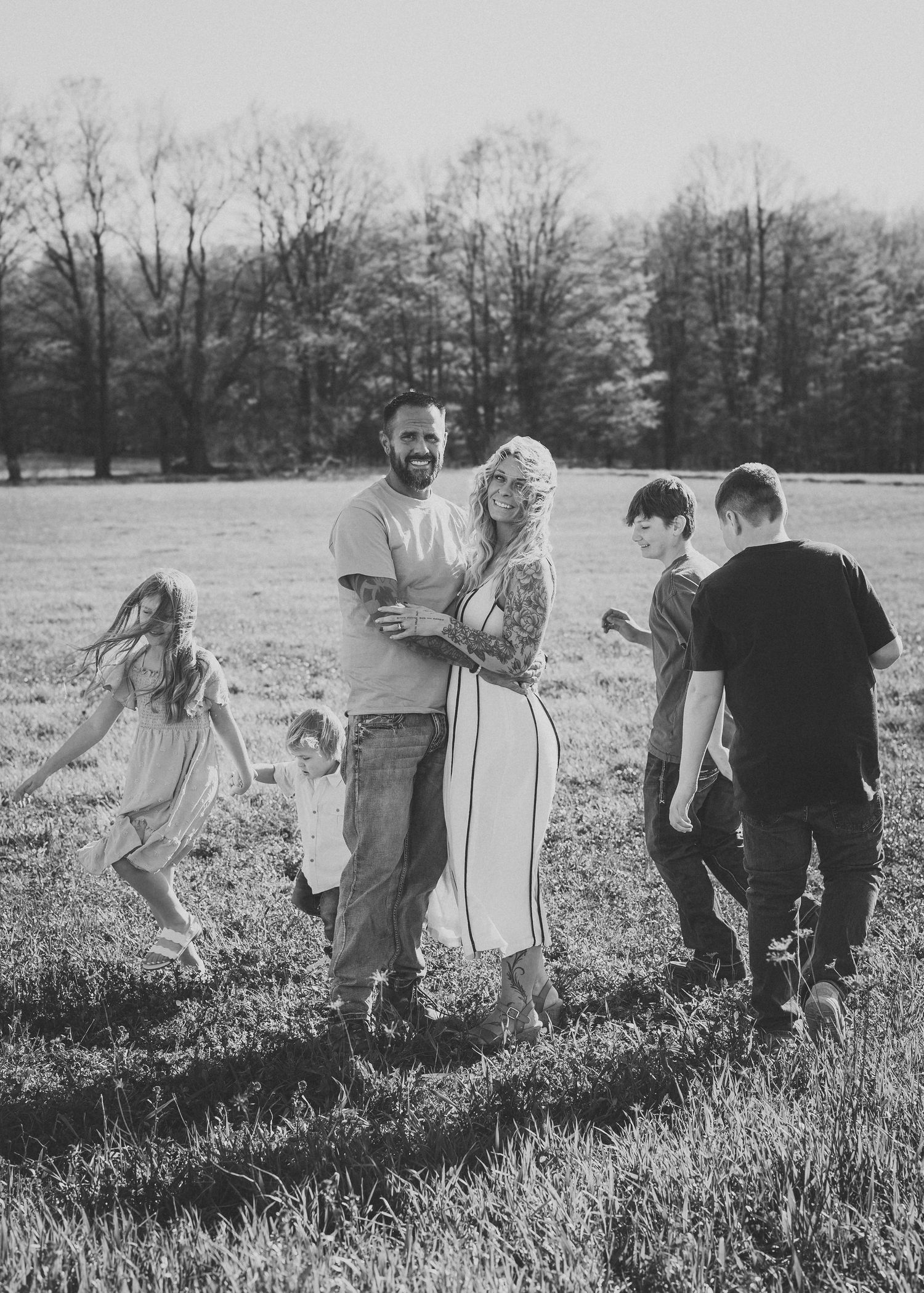 Black and white family photo of parents embracing in a field while four children play around them, with trees in background.