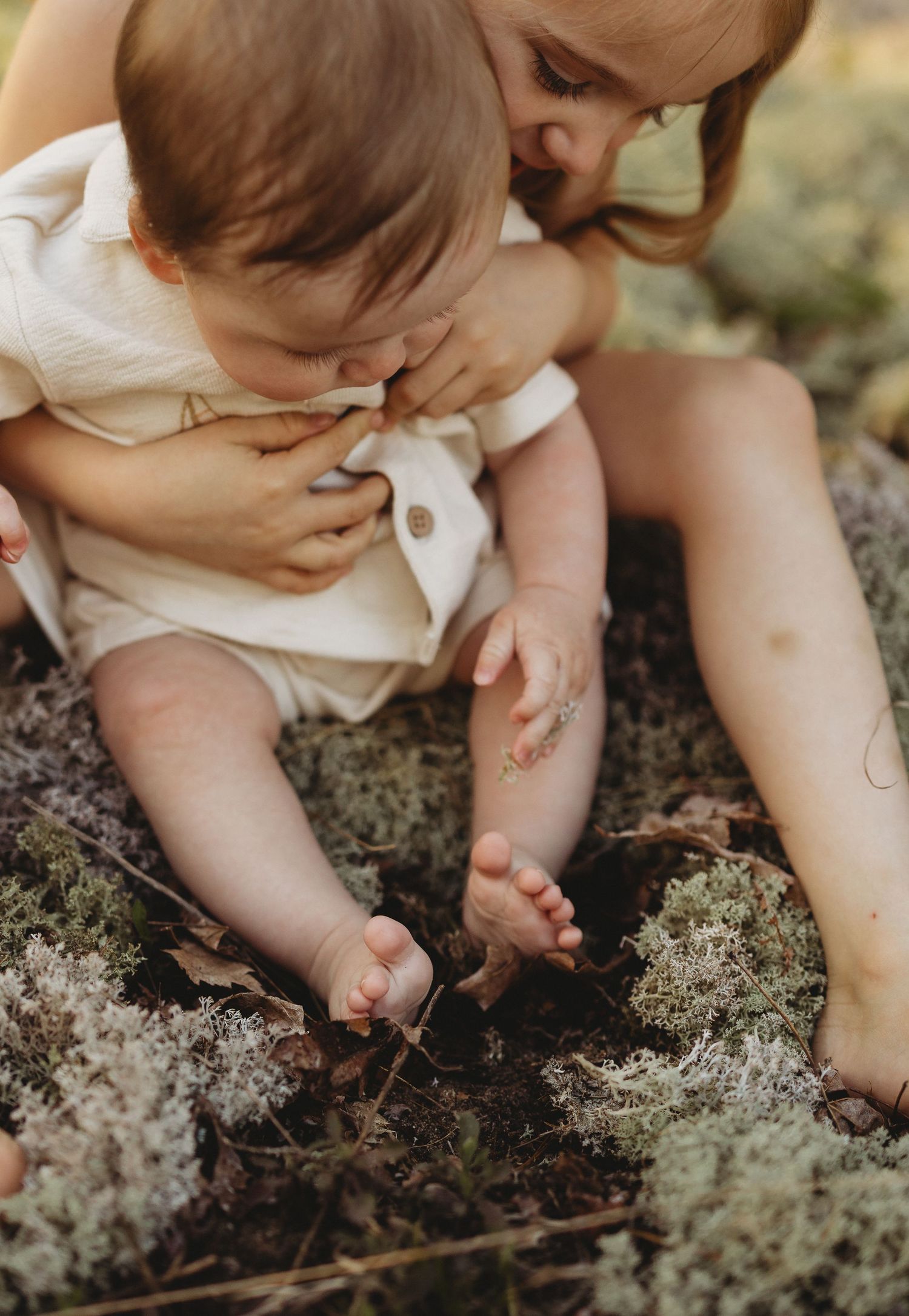 A baby explores moss and twigs on the ground while a mother watches closely in a natural outdoor setting.