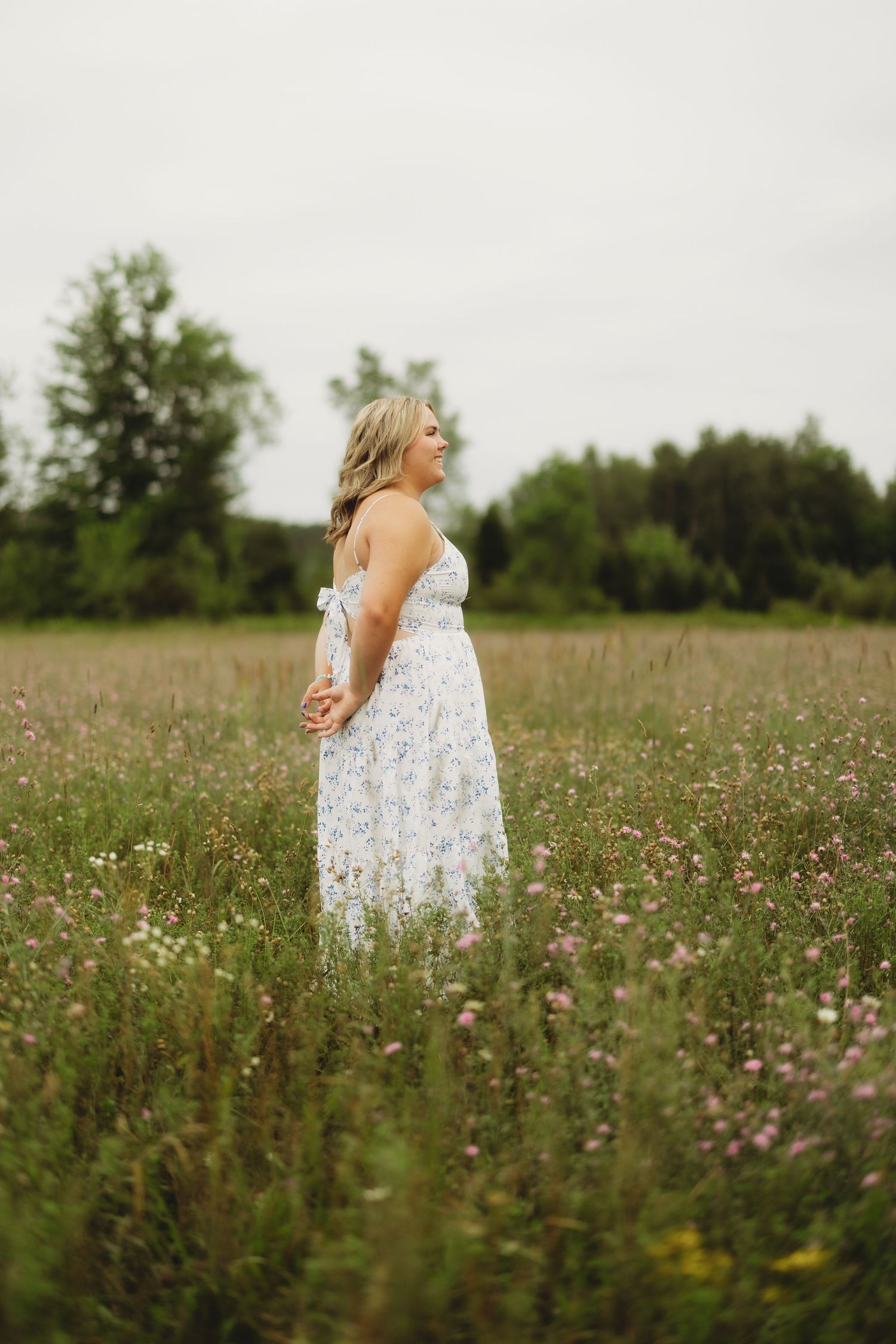 Pregnant woman in white floral dress standing in a wildflower meadow with green trees in the background.