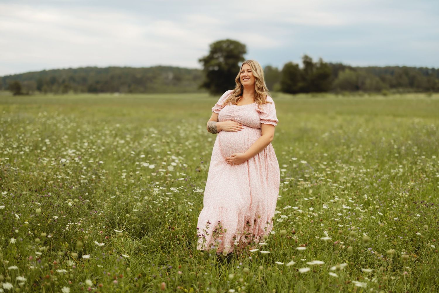 Pregnant woman in pink floral maxi dress standing in wildflower field during outdoor maternity photo session.