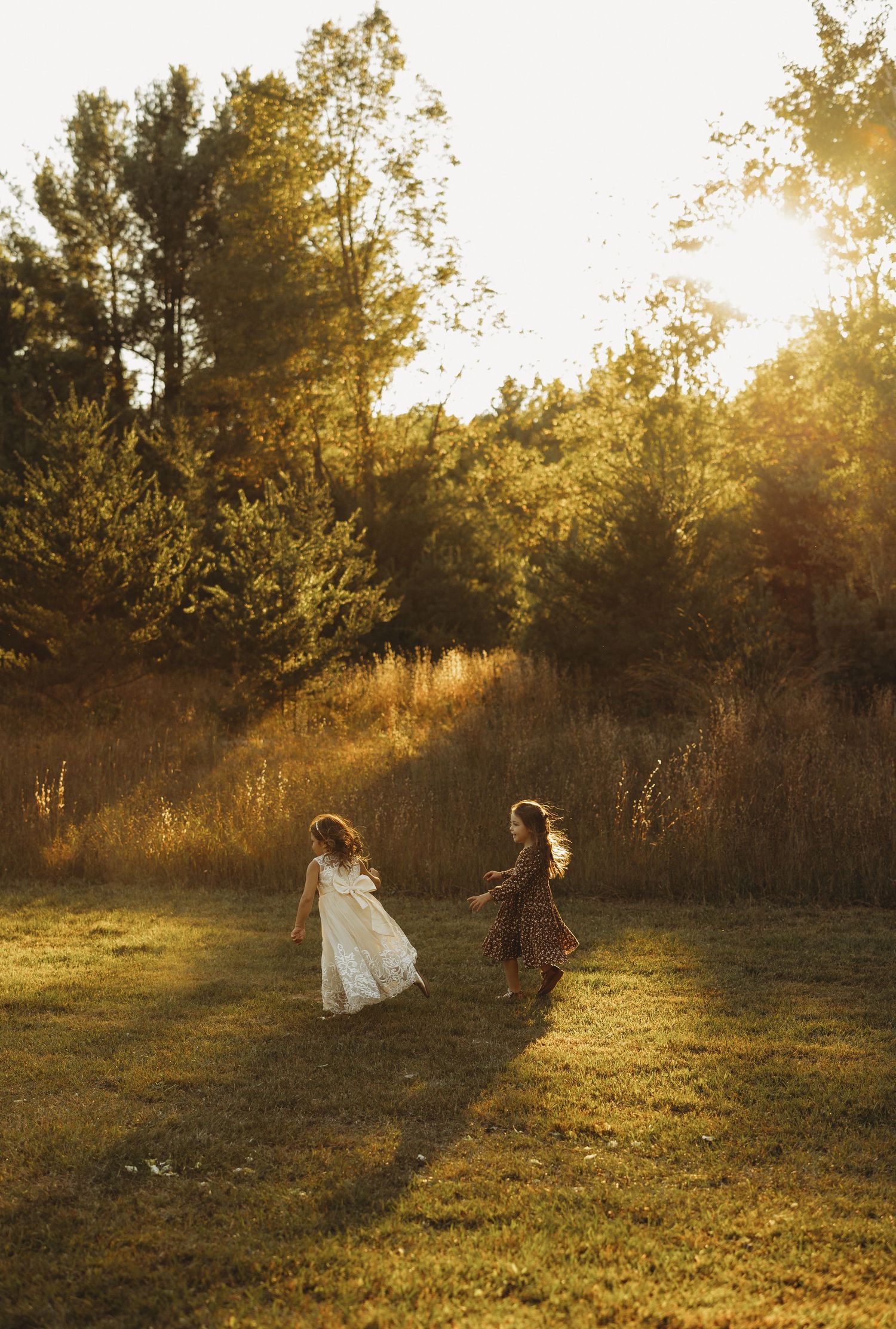 Two girls in dresses run and twirl across a grassy meadow at golden hour with sunlight glowing through evergreen trees.