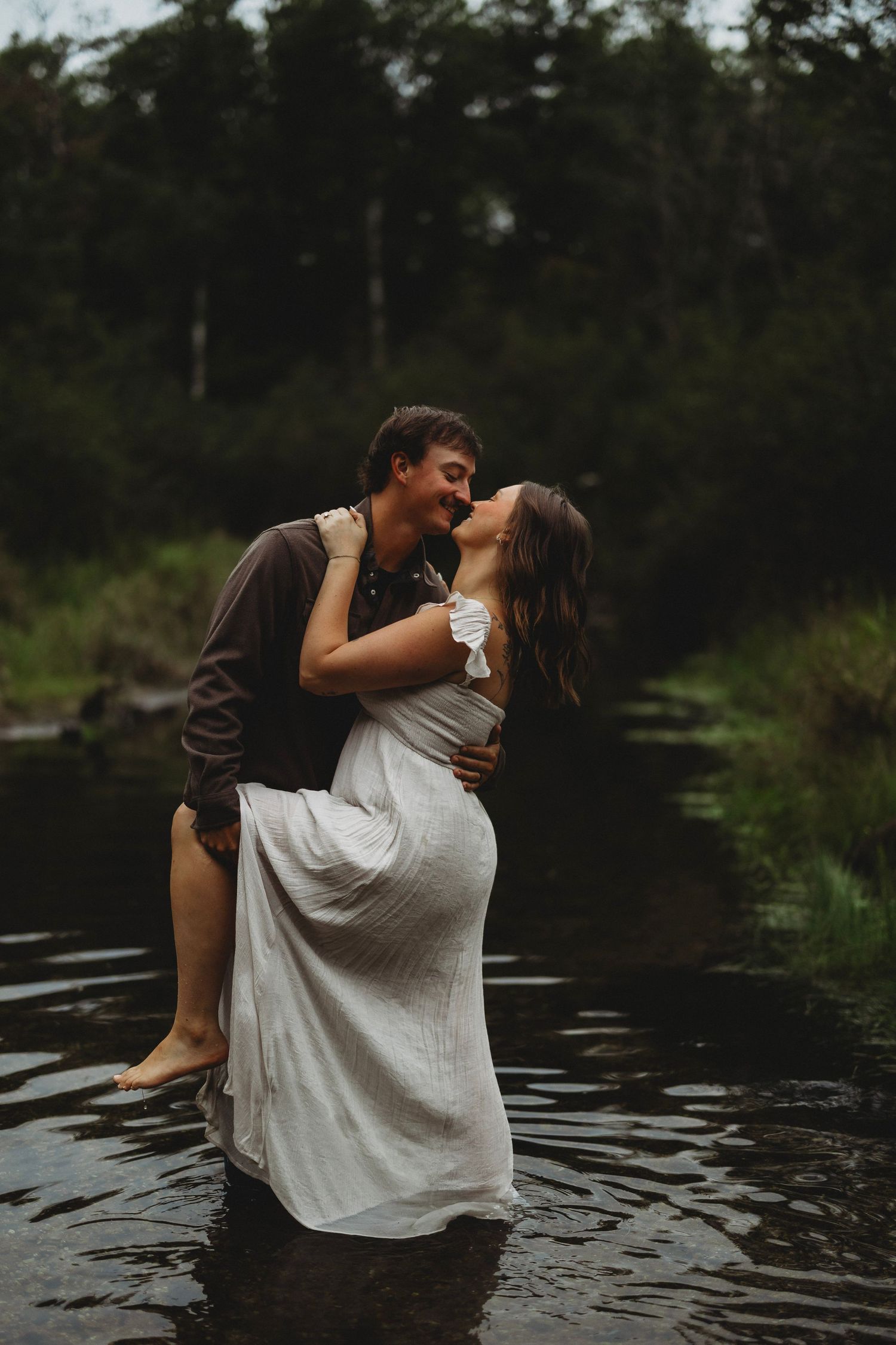 Couple in a romantic embrace, a barefoot man lifting a woman in a flowing white dress as they wade through a calm forest stream.