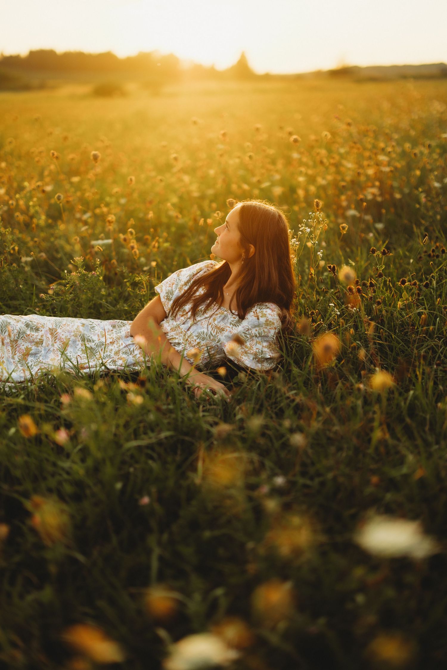 Woman in a floral dress lies back in a field of yellow wildflowers, bathed in warm golden sunset light.