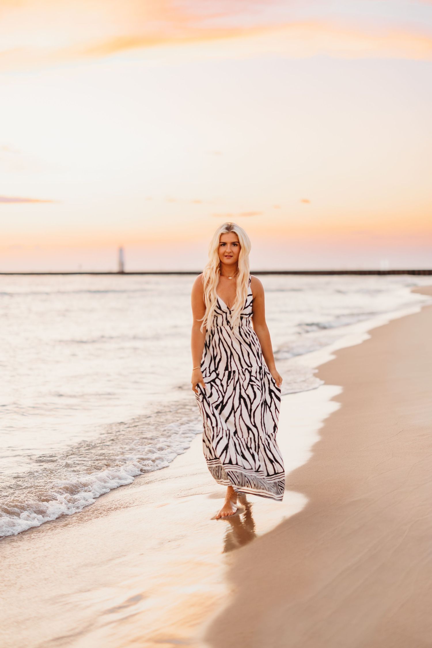 Blonde woman in a long zebra-print maxi dress walks barefoot along the shoreline at sunset with a pastel sky and distant lighthouse.