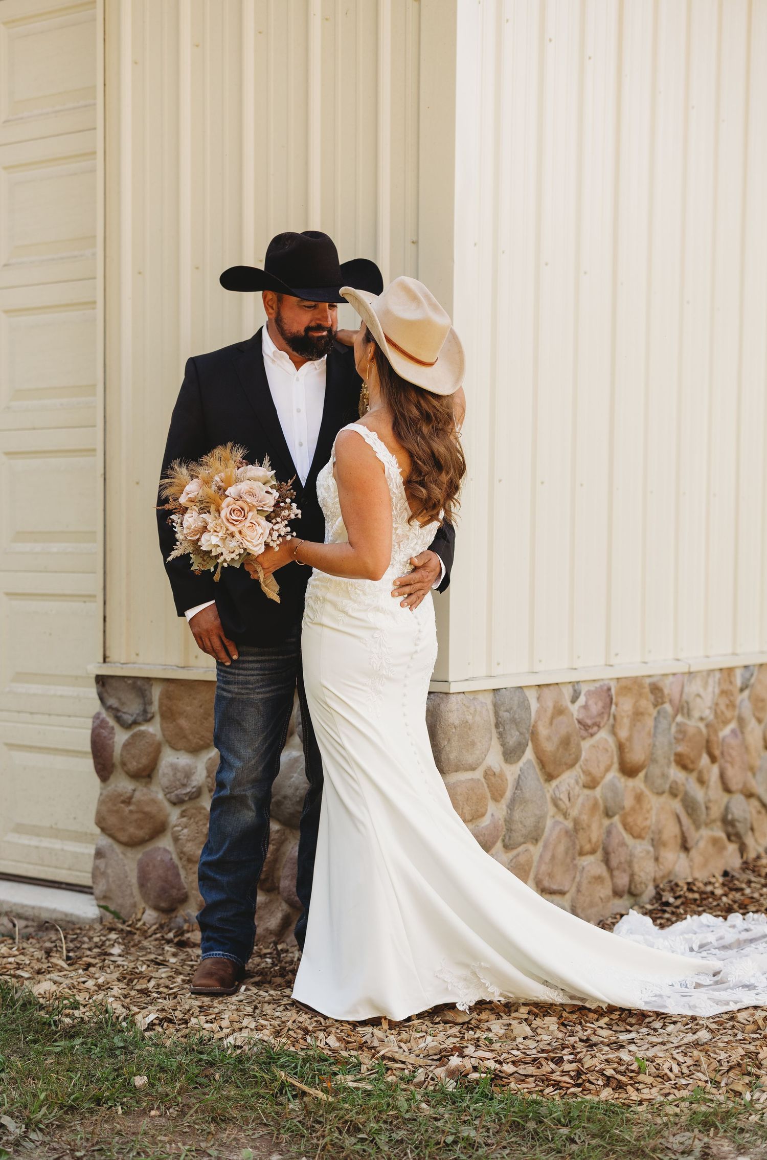 Bride in a fitted white gown and cowboy hat embraces a groom in a black hat and jacket while holding a dried floral bouquet.