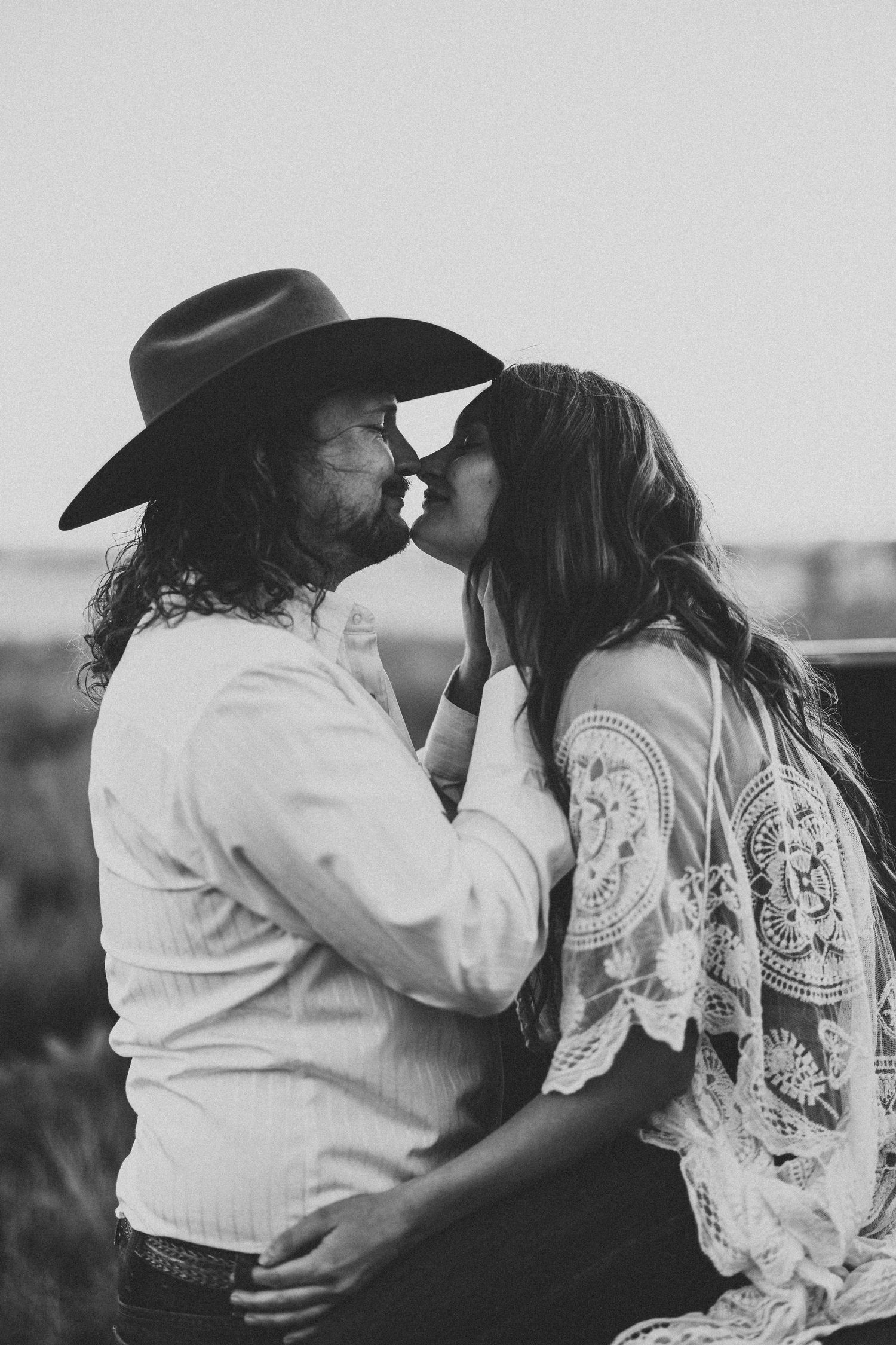 A romantic black and white photo of a couple sharing a tender kiss outdoors, wearing western and bohemian style clothing.