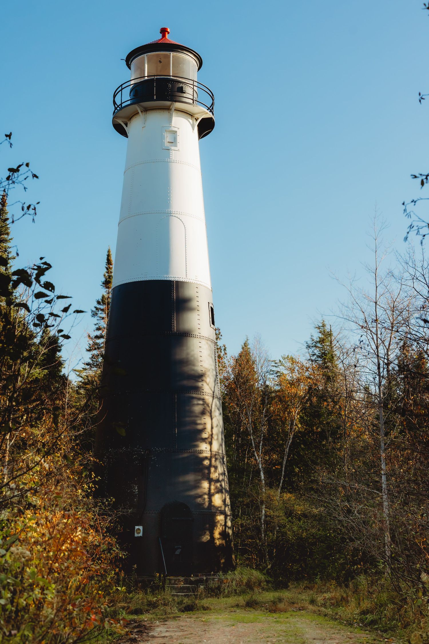 Tall black and white lighthouse tower stands against blue sky surrounded by fall foliage.