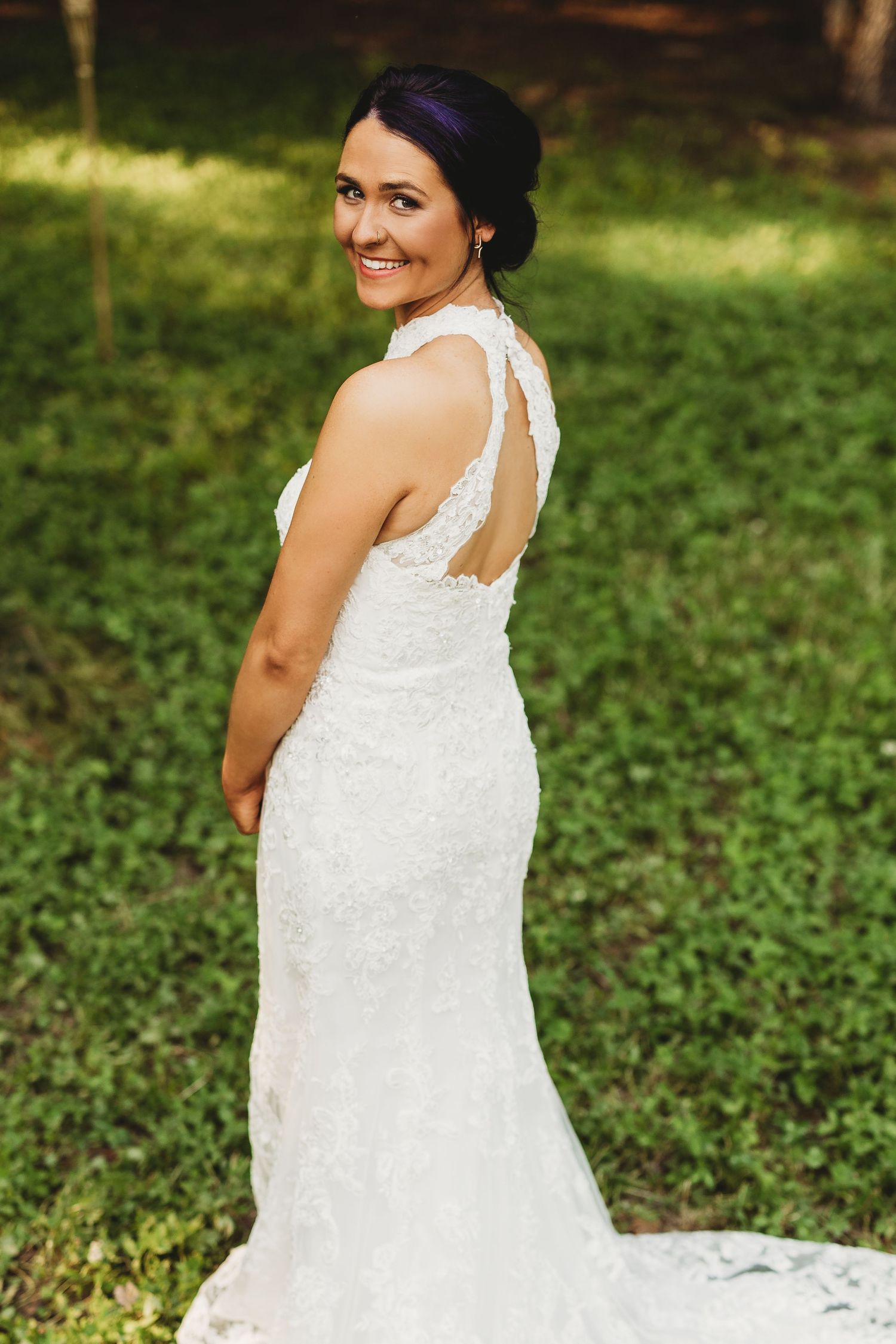 A bride in a flowing white lace wedding dress poses outdoors in a garden setting with natural lighting.