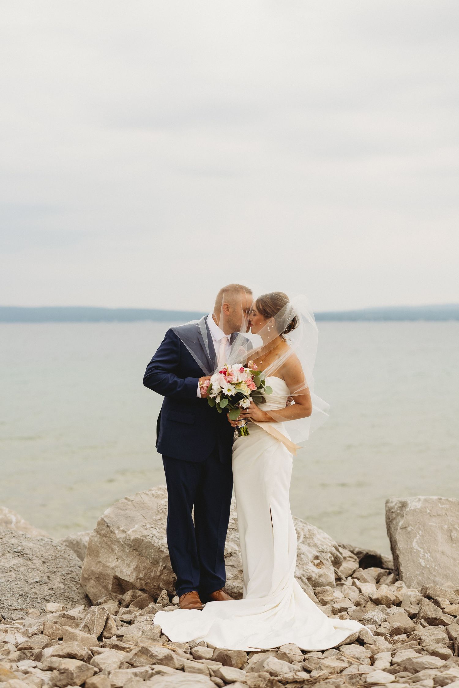 Bride and groom kissing on rocky shoreline, bride in white gown holding bouquet, groom in navy suit, ocean backdrop.