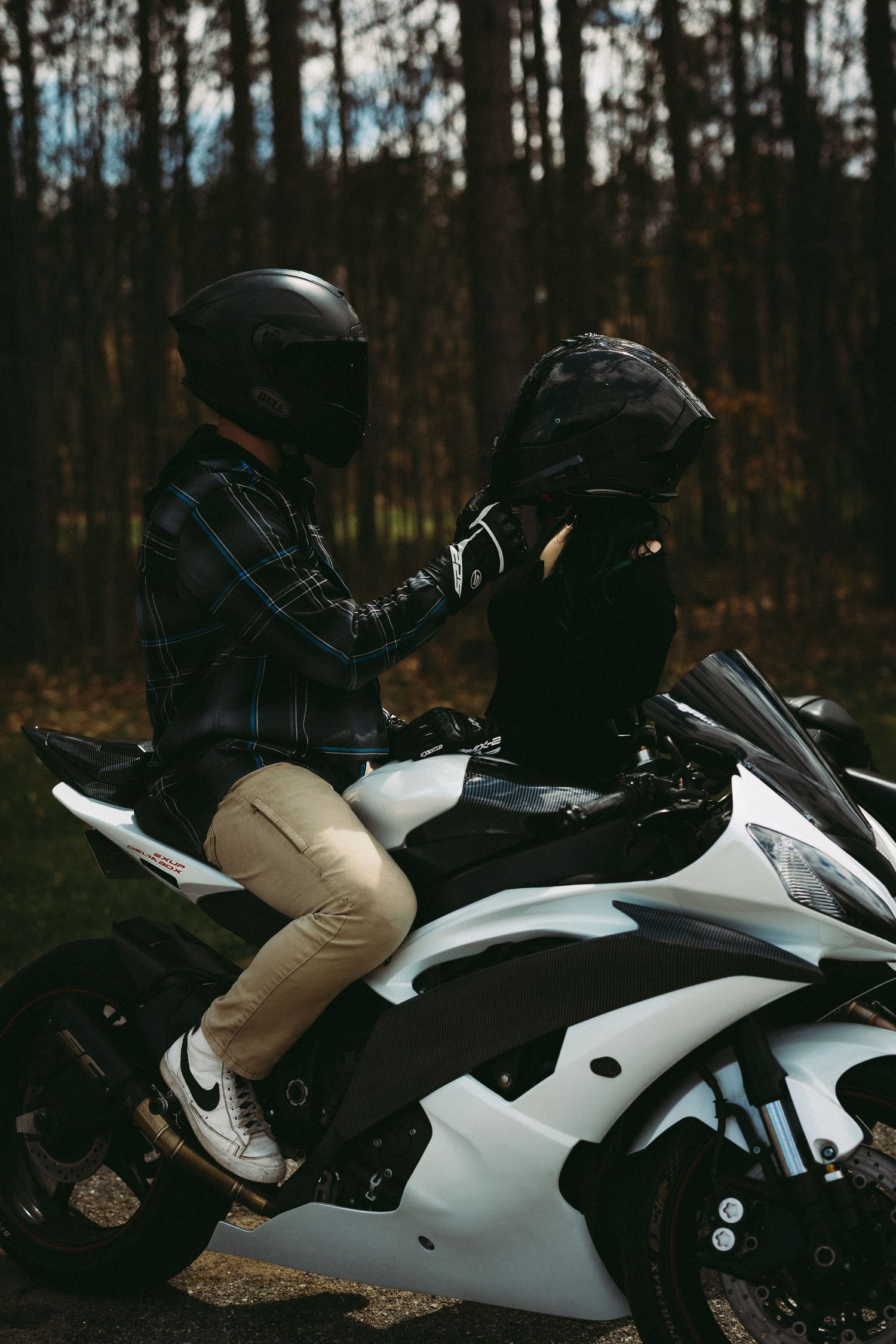 Two motorcyclists in black helmets sitting on a white sport bike in a forest setting.