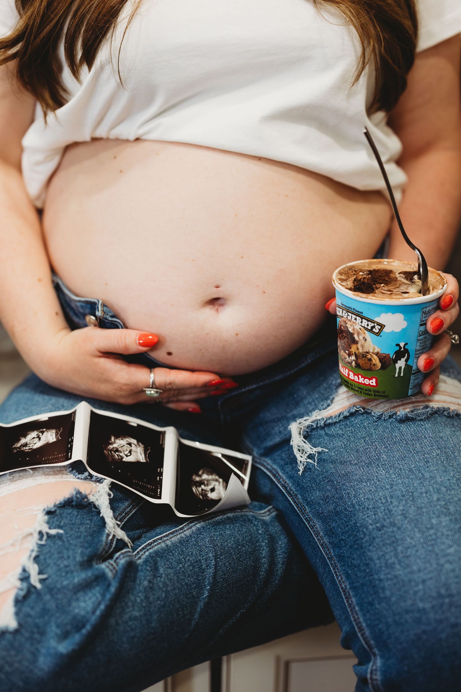 Pregnant woman in ripped jeans holding Ben & Jerry's ice cream with ultrasound photos on her lap.