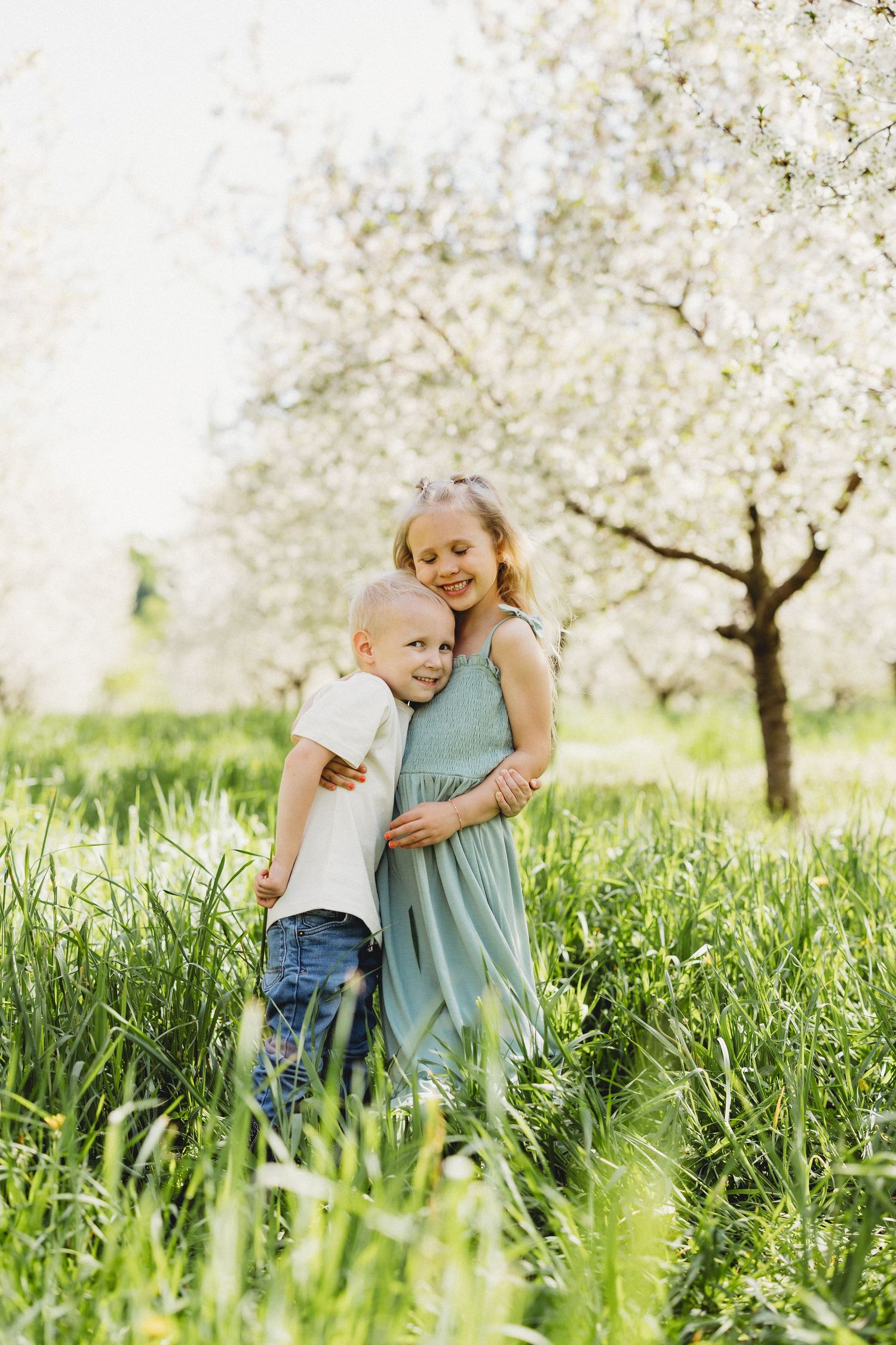 A young girl in a mint dress hugs her little brother in a sunny blooming orchard with tall green grass.