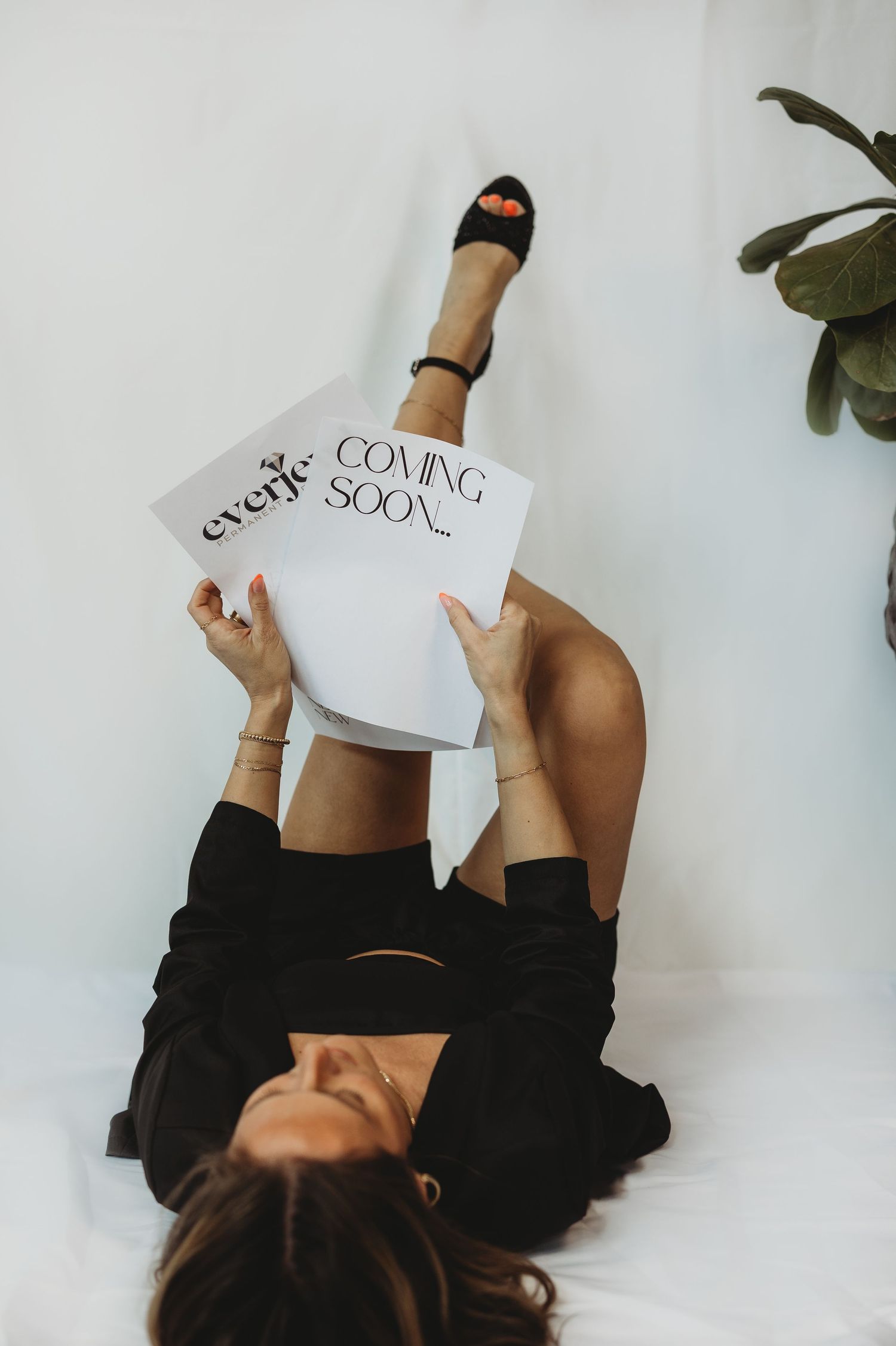 Woman lying down holding a 'Coming Soon' sign, wearing black outfit and heels, with a plant in the background.