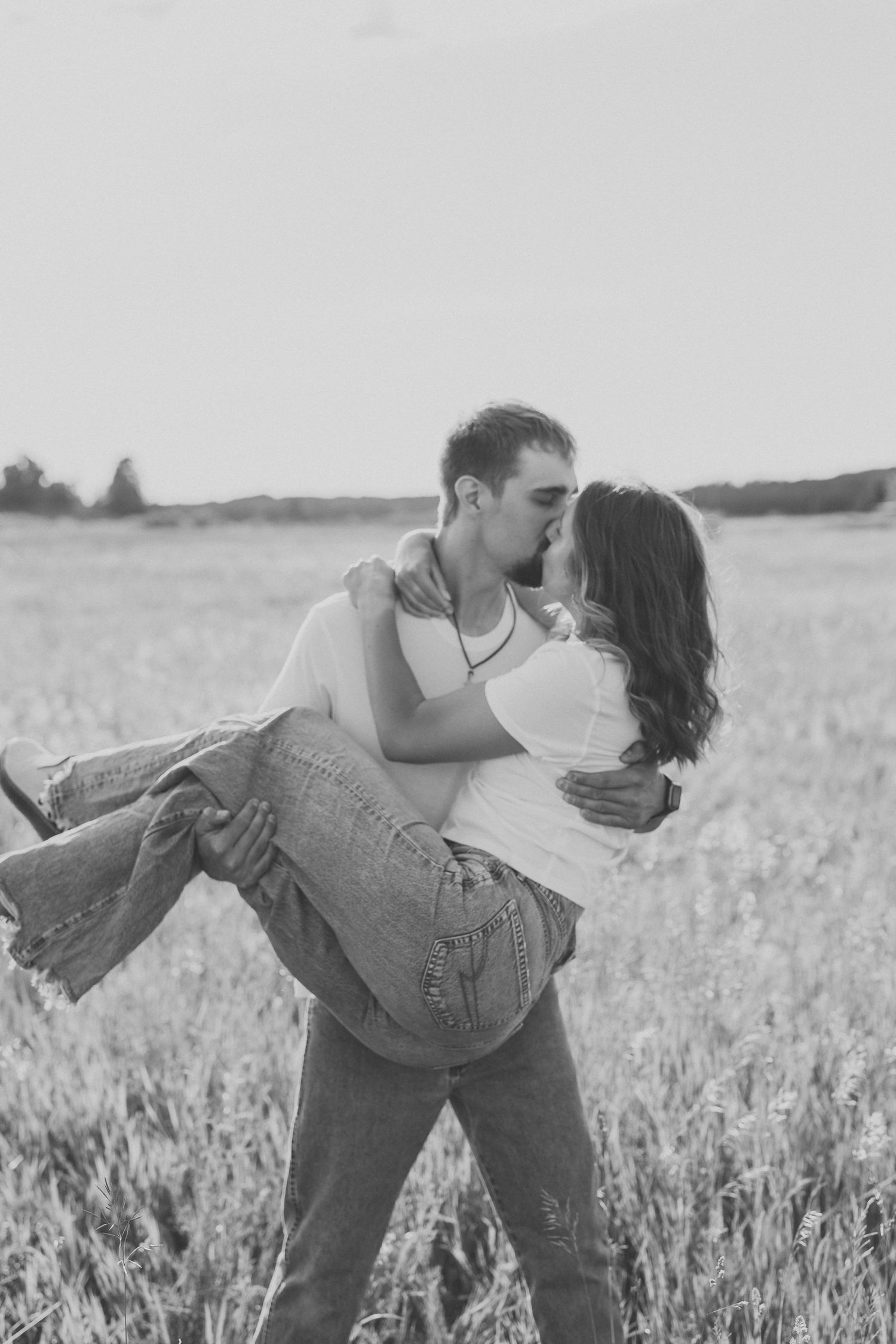 Black and white photo of a romantic couple kissing in a field, man lifting woman in his arms.
