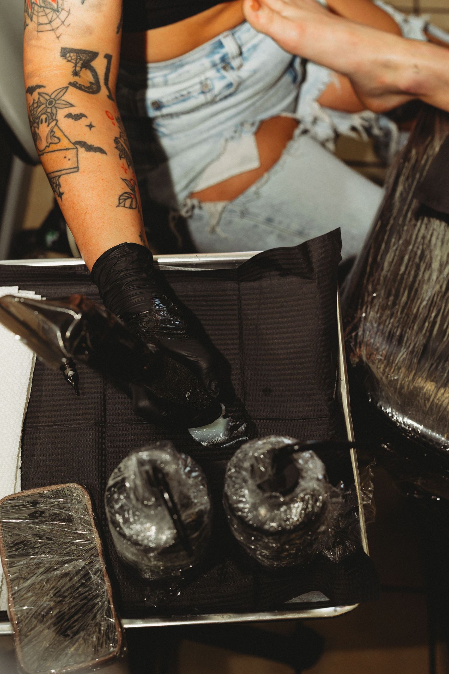 Tattooed hairstylist in black gloves applying dark hair dye from mixing bowl during salon coloring service.