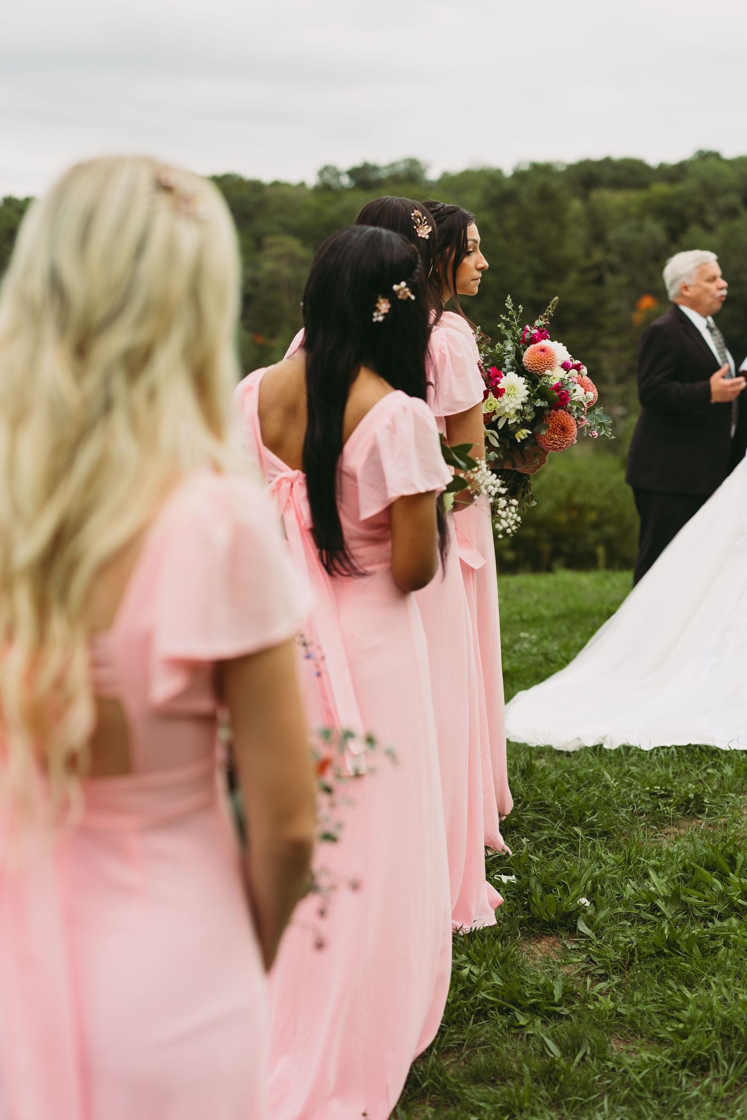 Bridesmaids in flowing pink dresses stand together on a grassy hill during an outdoor wedding.