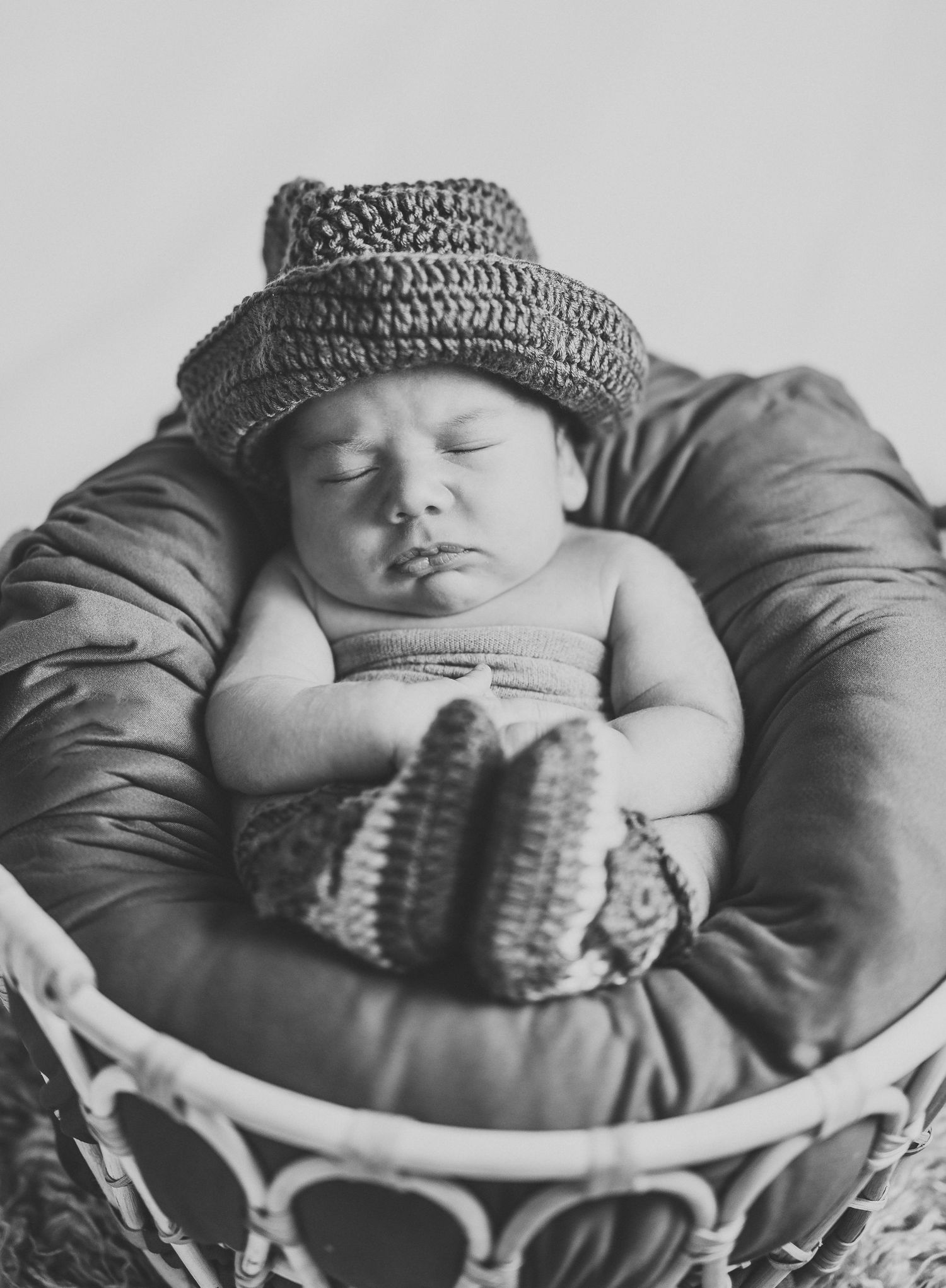 Black and white portrait of sleeping newborn wearing knitted hat and mittens on soft cushion.