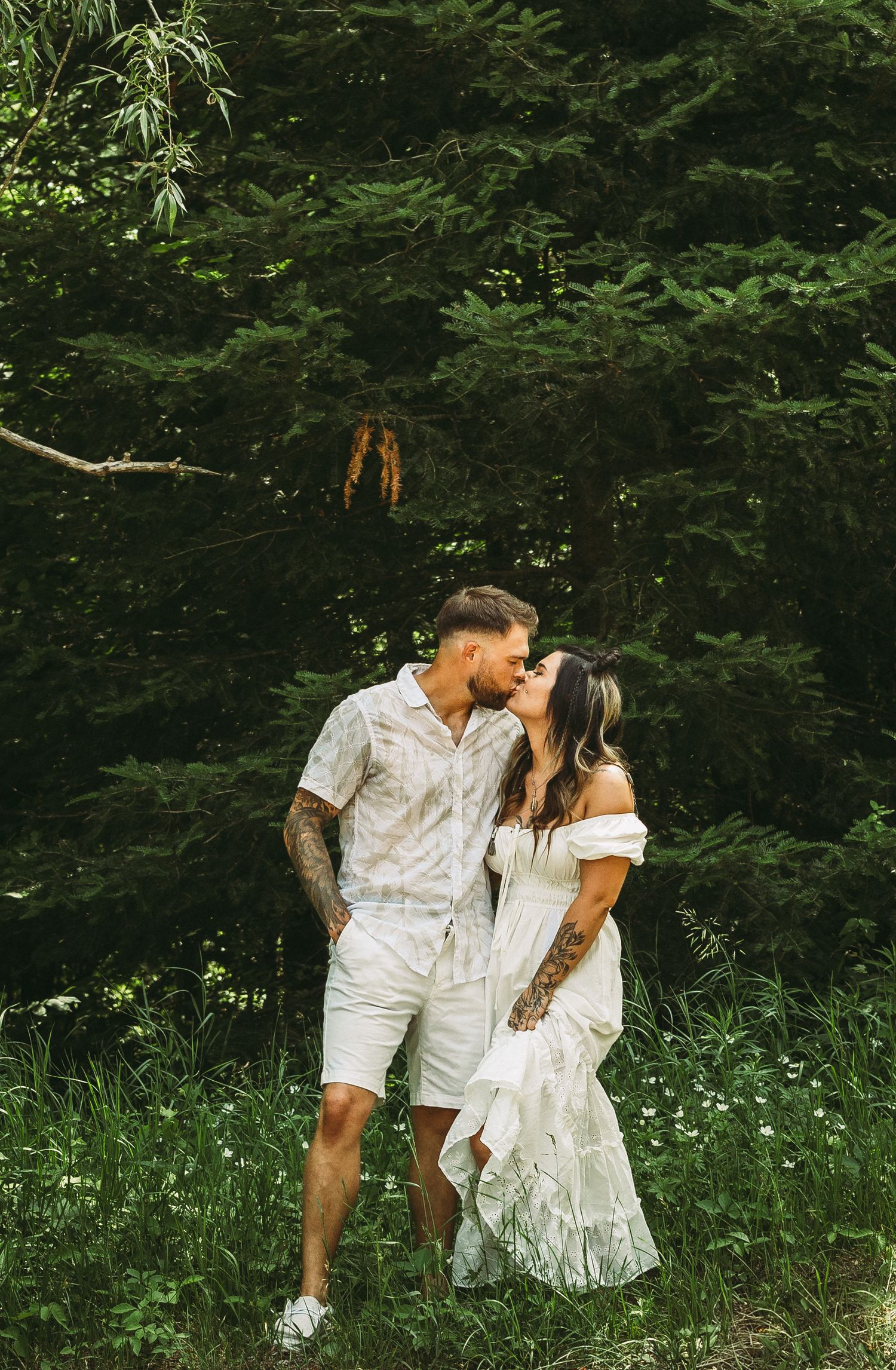 Romantic couple in white outfits embrace and share intimate moments in a lush green meadow with wildflowers.