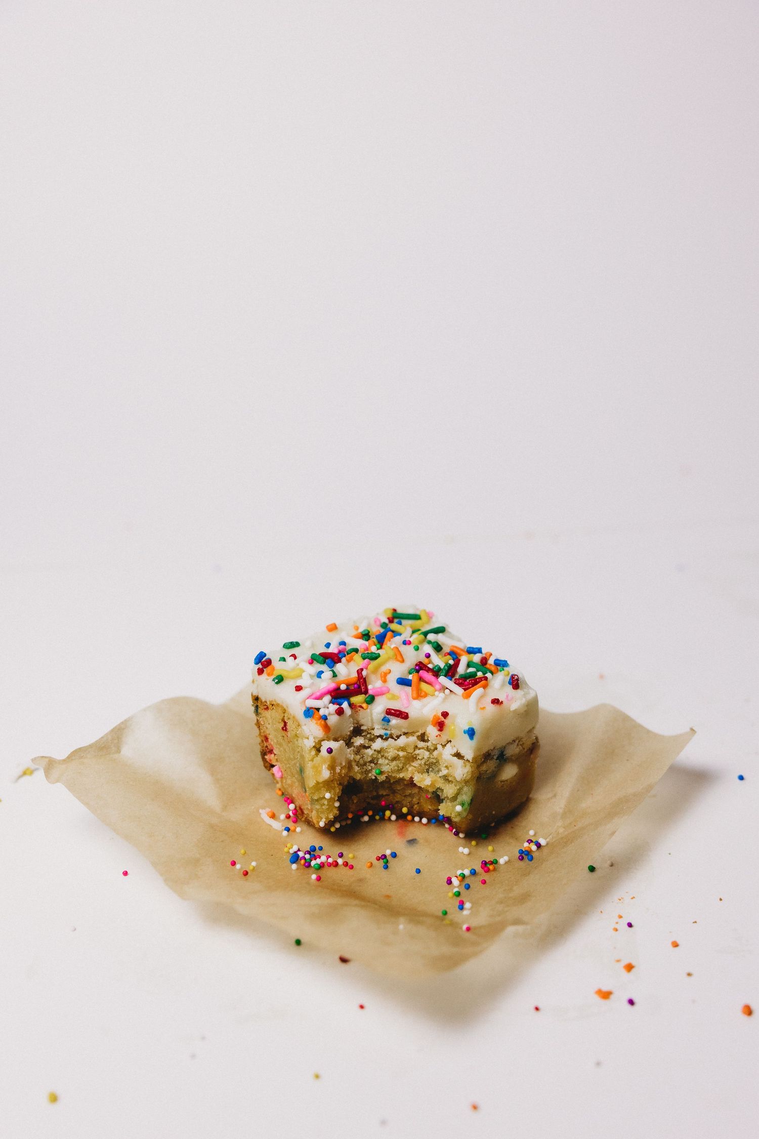 Single frosted vanilla cake bite with colorful sprinkles on parchment paper against a white background.