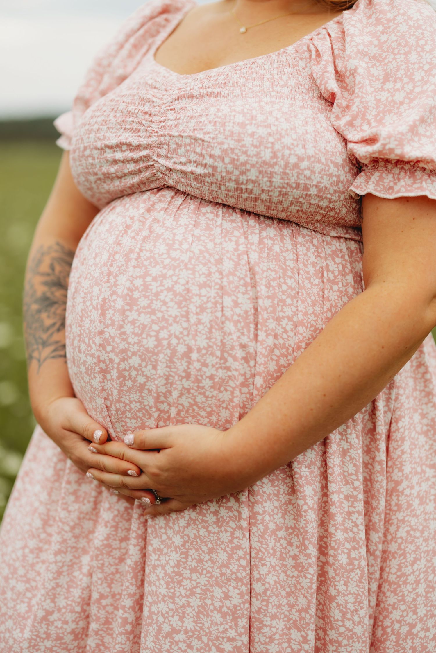 Pregnant woman in pink floral dress cradling baby bump outdoors, showing tattoo on arm.