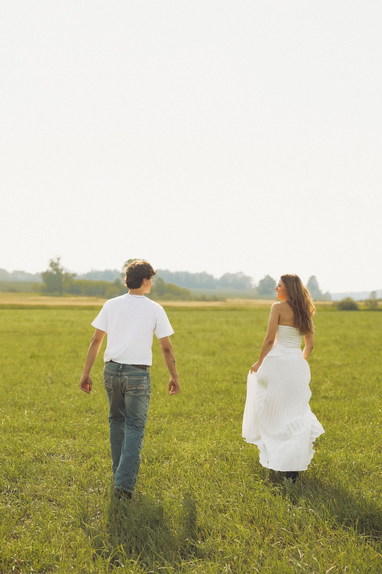Couple walking through a sunny green meadow, man in jeans and woman in white dress strolling together.