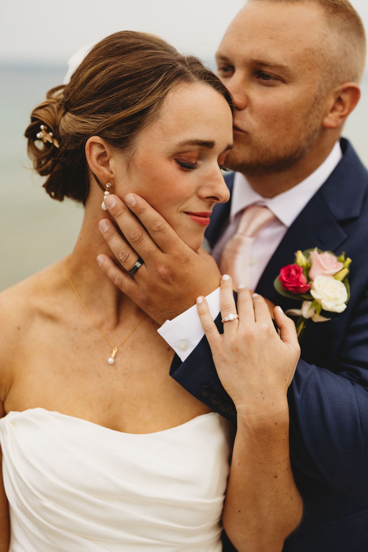 Groom in navy suit kisses bride wearing white dress and wedding rings in romantic outdoor ceremony photo.
