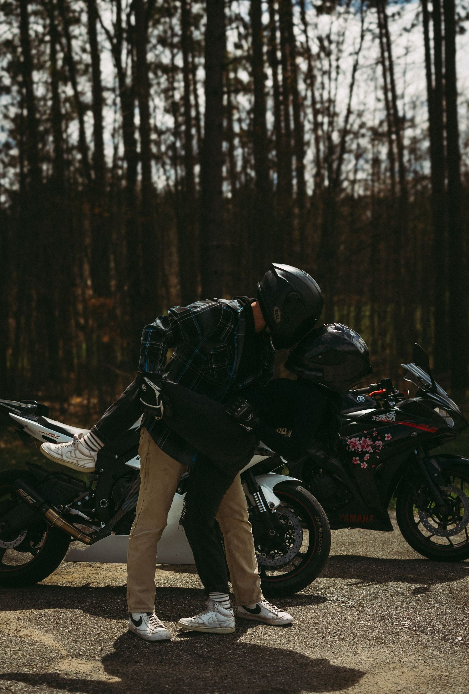 A motorcyclist in a helmet and jacket leans over a sport motorcycle parked on a forest road surrounded by tall trees.