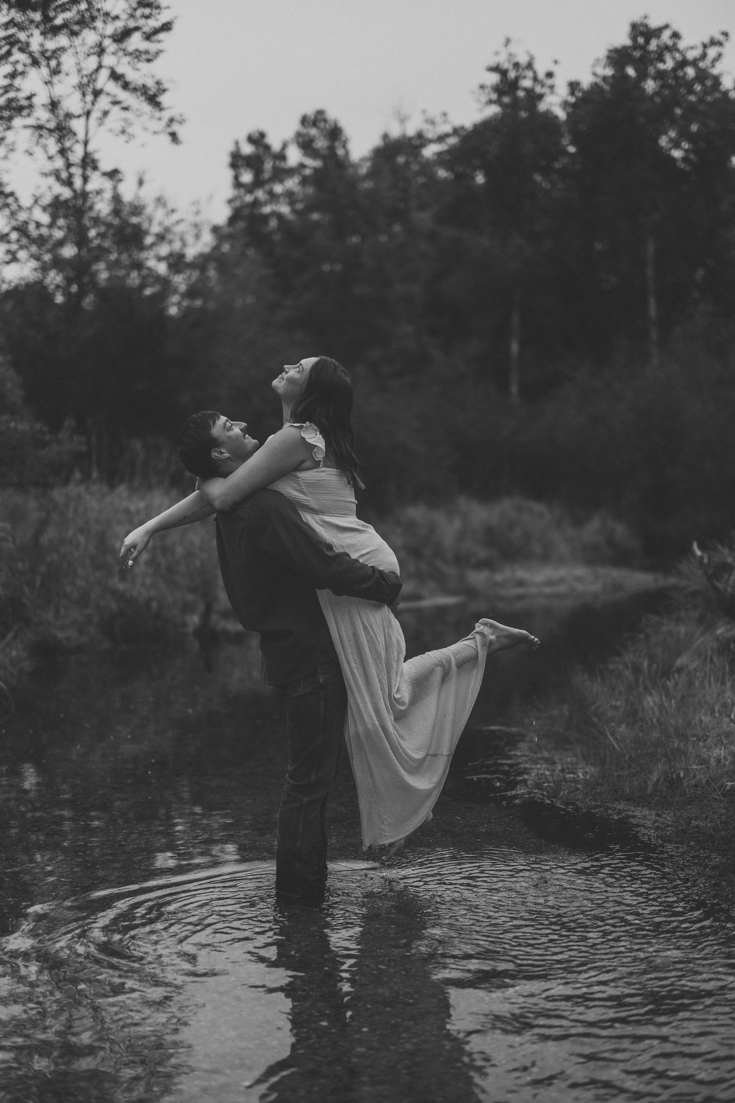 Black and white photo of a man lifting a woman in a flowing dress as they stand in a shallow stream surrounded by trees.