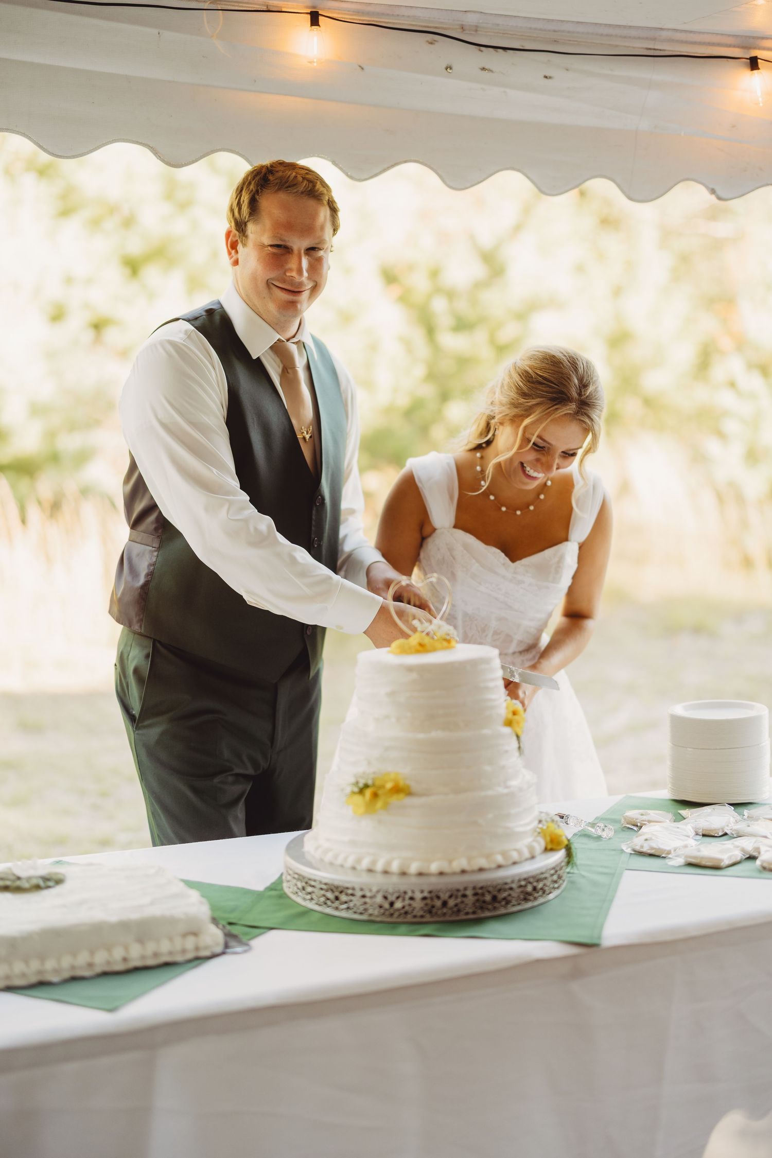 Smiling bride and groom cutting their three-tier white wedding cake with yellow flowers at an outdoor tented reception.