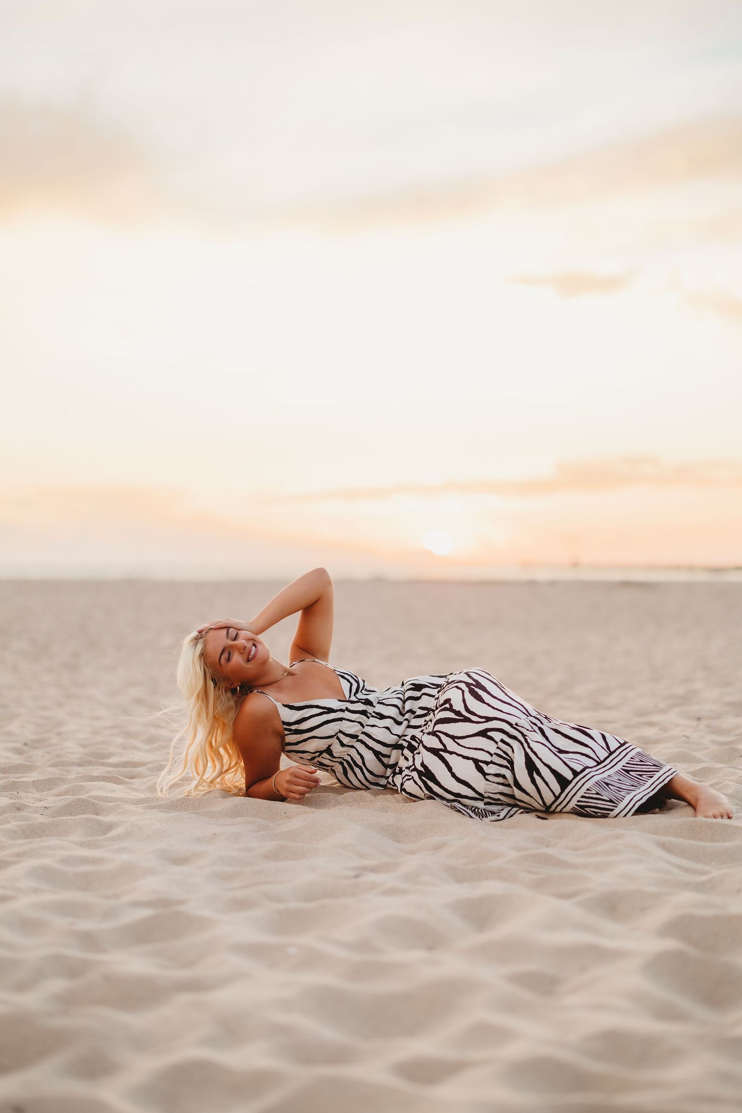 Blonde woman in a zebra-print maxi dress reclines and smiles on a sandy beach with a soft pastel sunset sky glowing behind her.