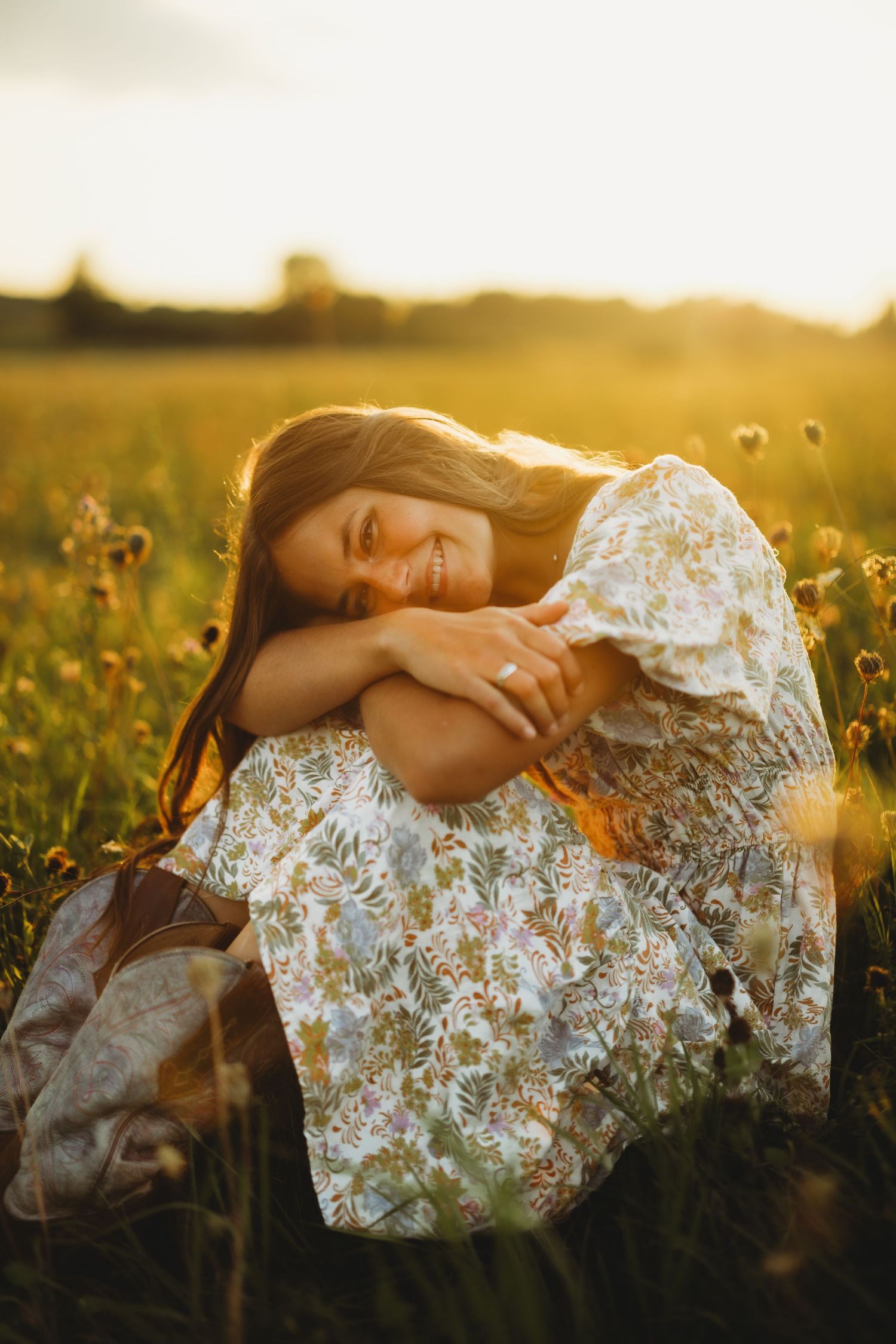 Smiling woman in a floral dress hugs her knees while sitting in a golden wildflower field at sunset.