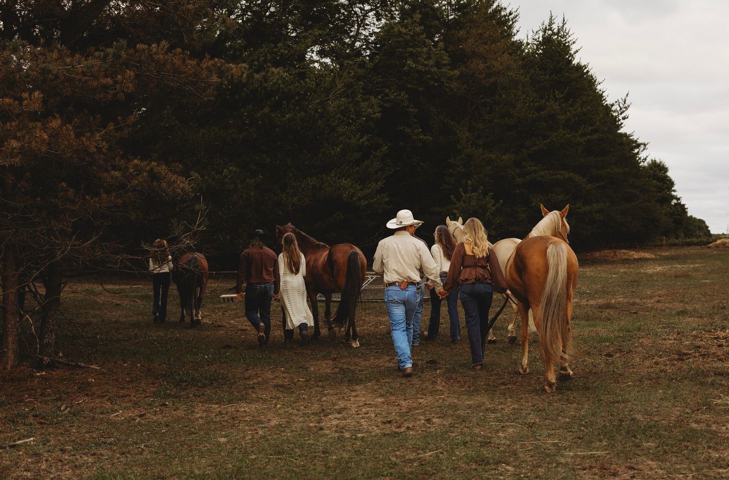 Group of people in cowboy hats and western wear leading palomino and brown horses across a grassy field toward a dense tree line.