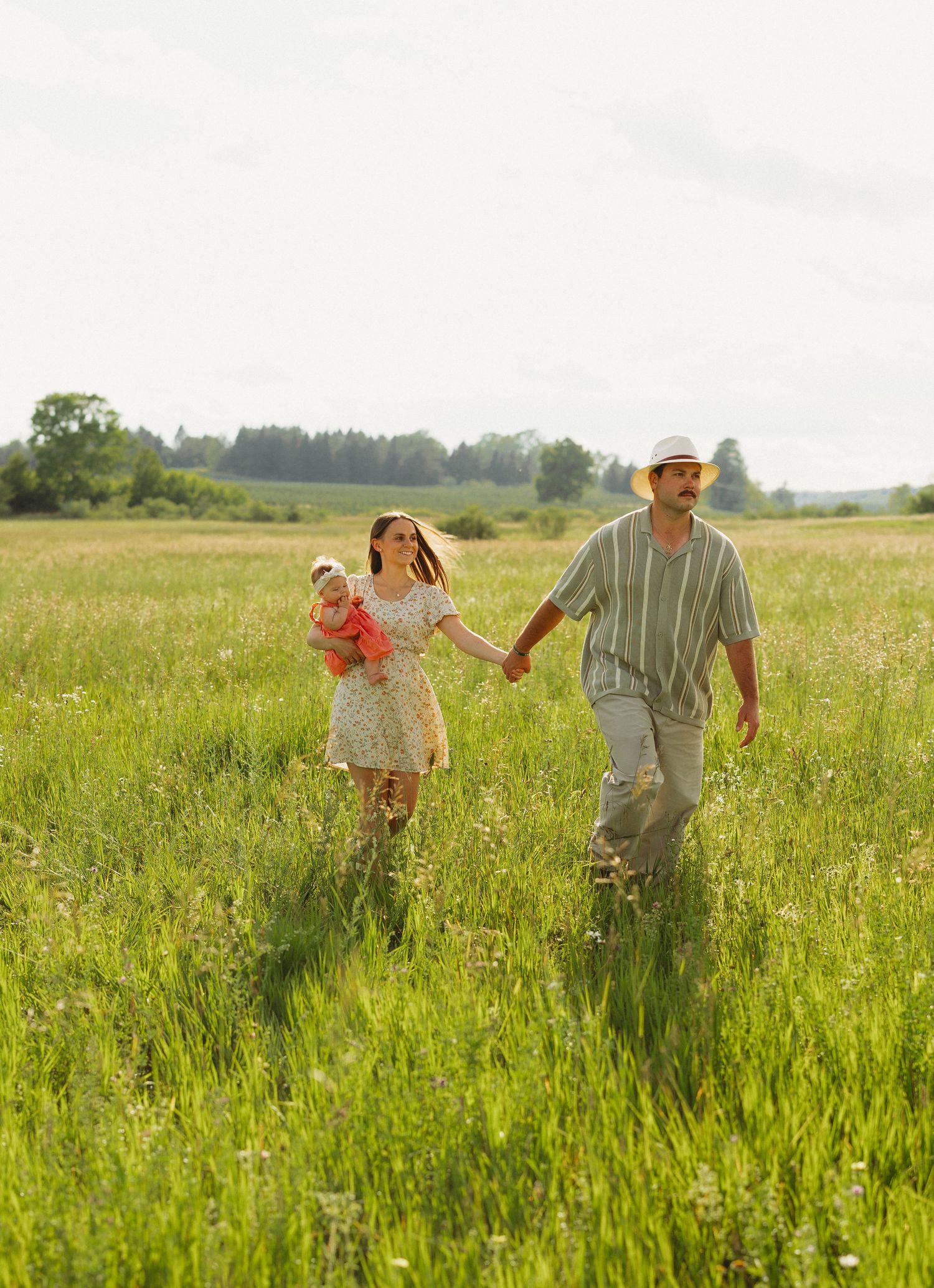 A family walks hand-in-hand through a lush green meadow, with a woman carrying a baby and a man in a straw hat.