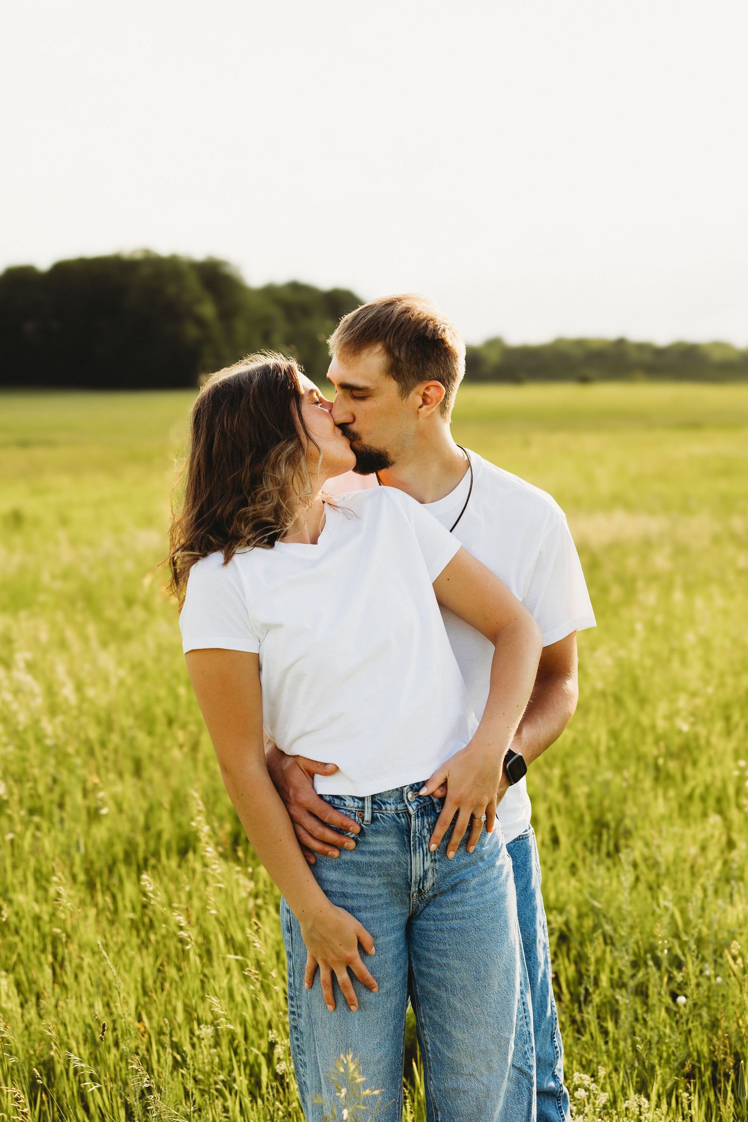 A couple in matching white t-shirts and jeans share a romantic kiss in a sunny green field.