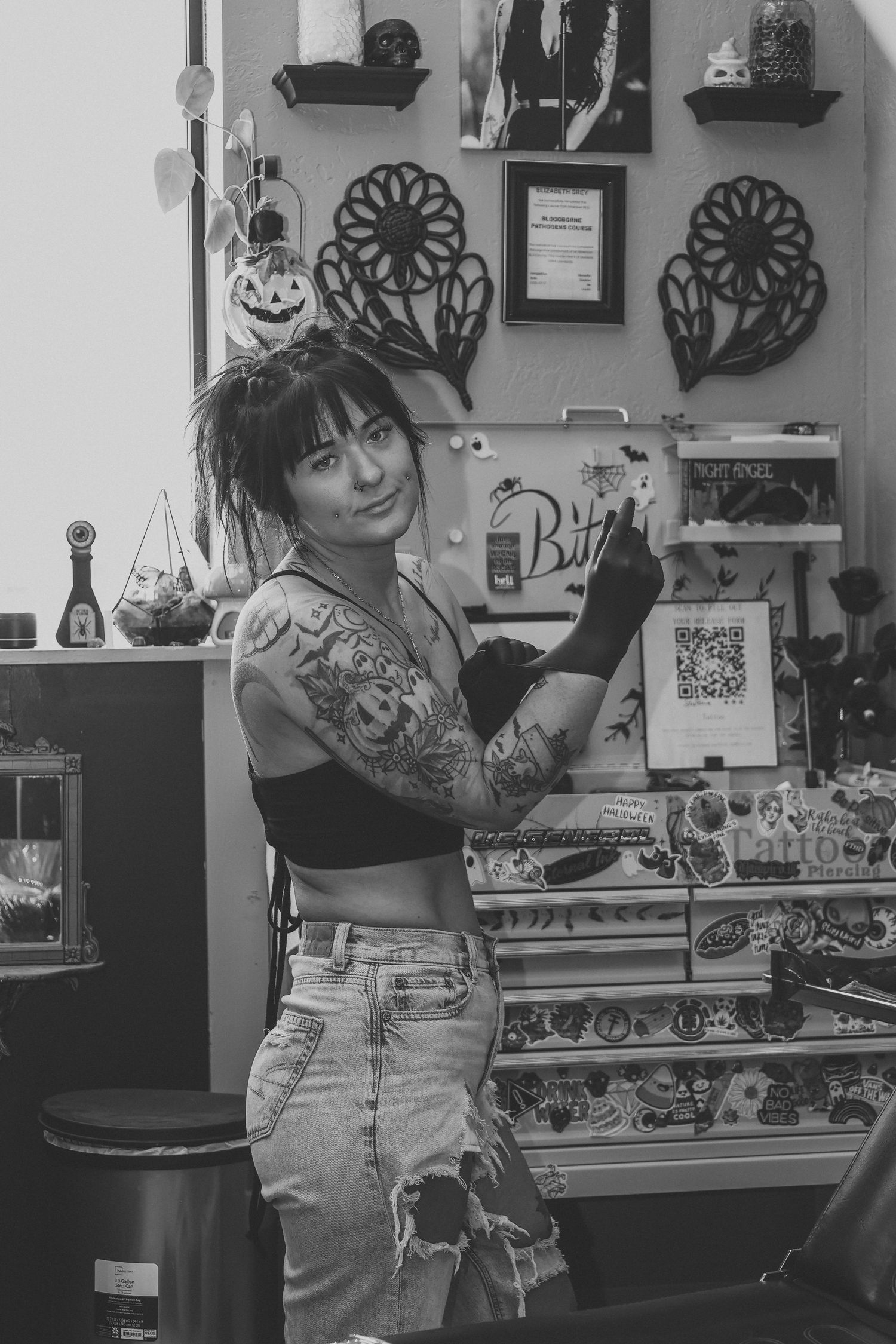 Tattooed female tattoo artist in black crop top and jeans posing in her decorated tattoo studio.