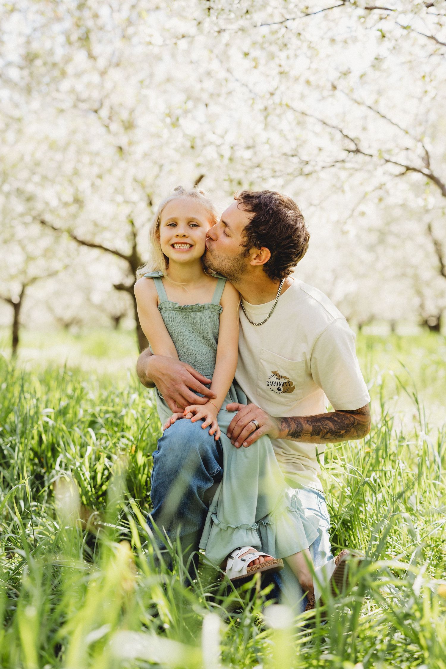 A father kisses his smiling blonde daughter on the cheek in a lush green field with white blossoming trees.