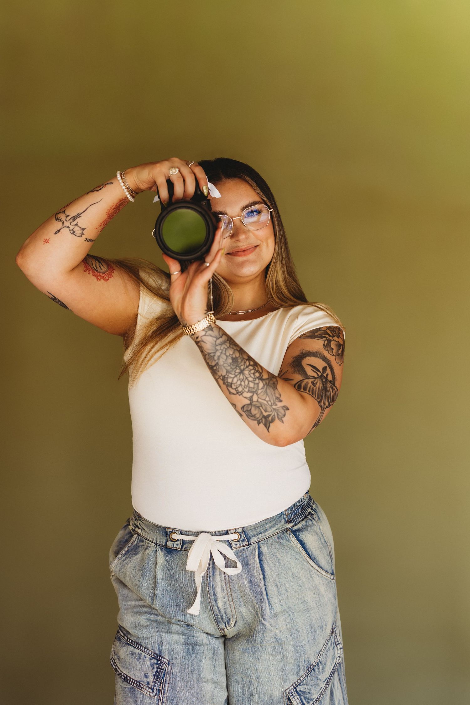 Tattooed woman in white top and jeans holds DSLR camera up to face, smiling, against olive green background.