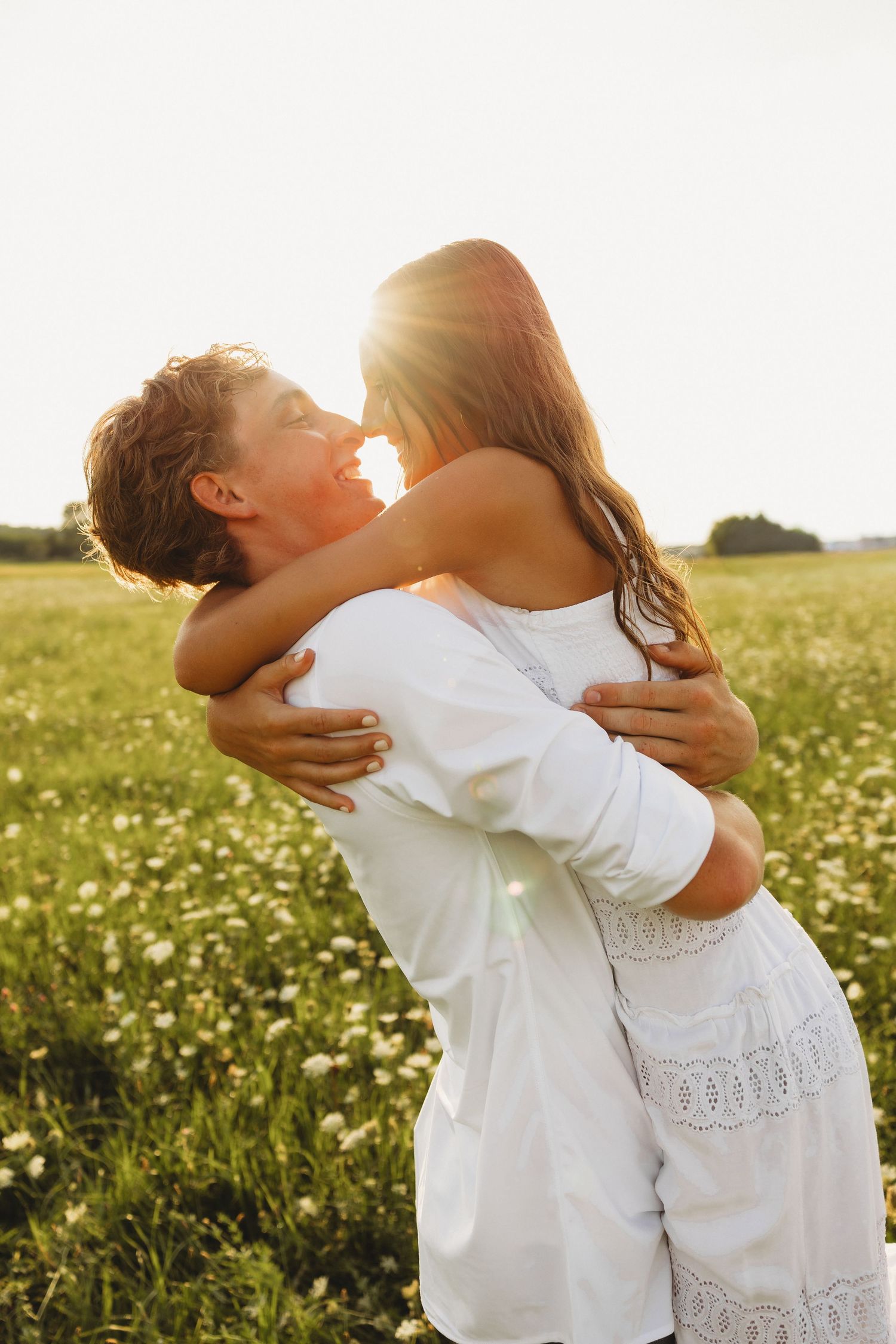 A couple in white embraces in a sunlit flower field, sharing a romantic moment with golden backlight.
