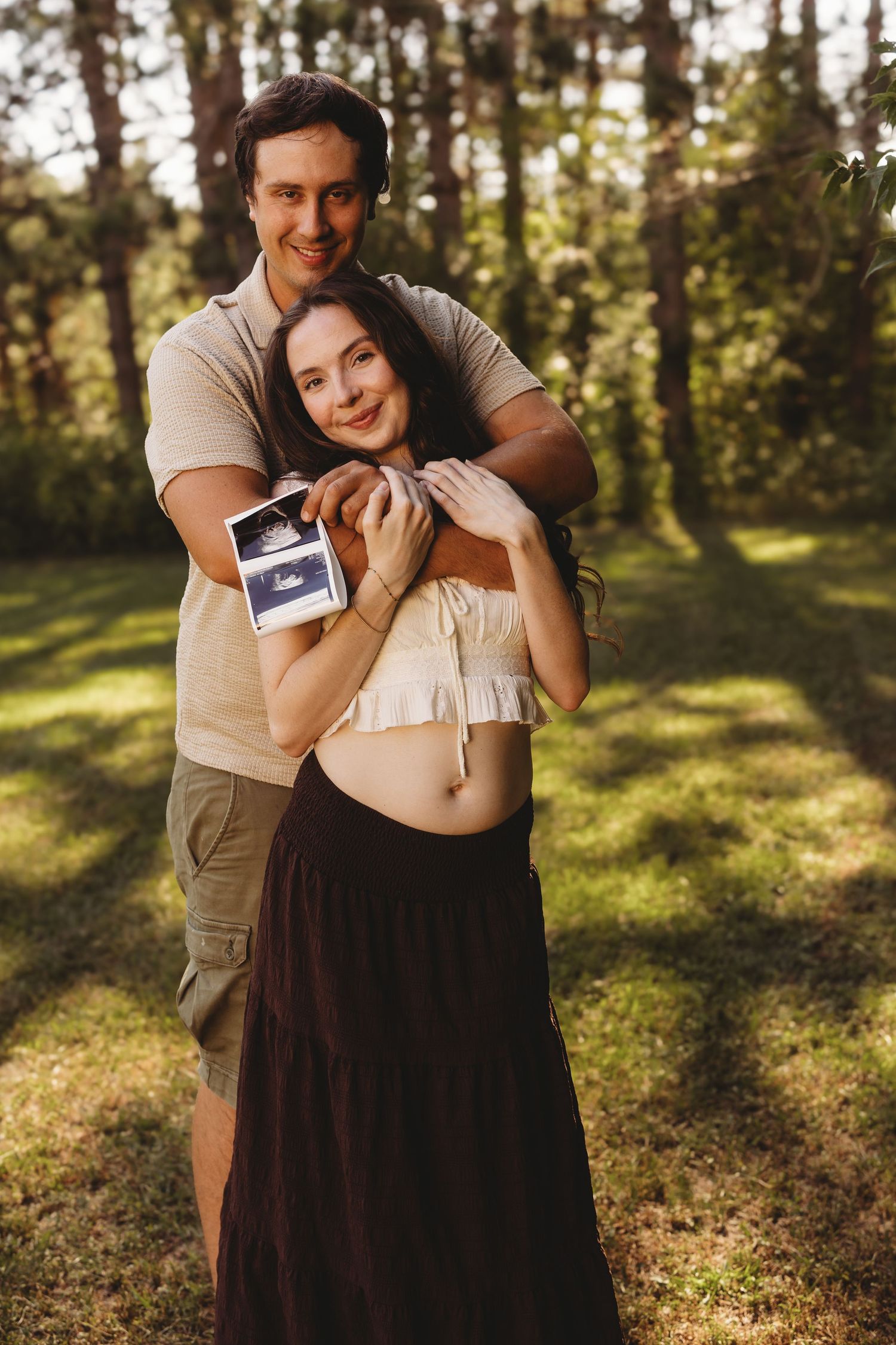 Happy couple embracing outdoors holding ultrasound photos, pregnant woman showing baby bump in sunny forest setting.