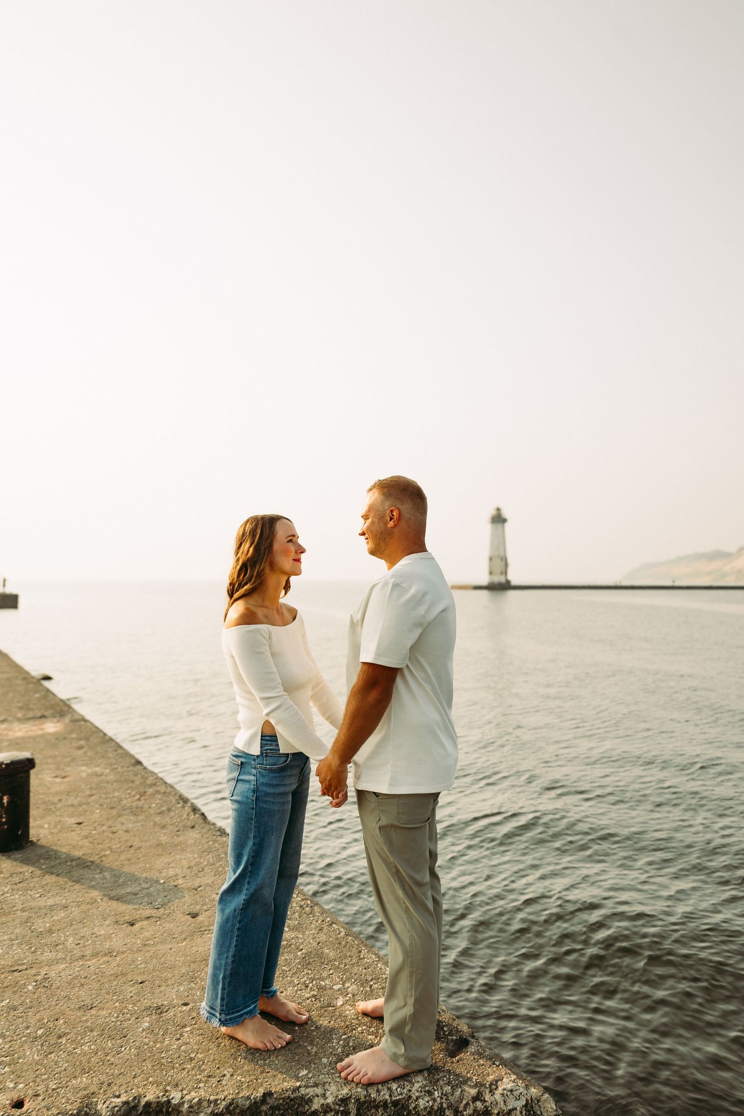 Couple holding hands barefoot on a waterfront pier at sunset with a lighthouse in the background.