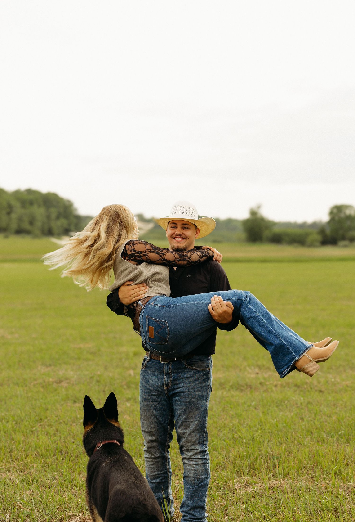Man in cowboy hat lifting smiling woman in field with German Shepherd dog standing nearby.