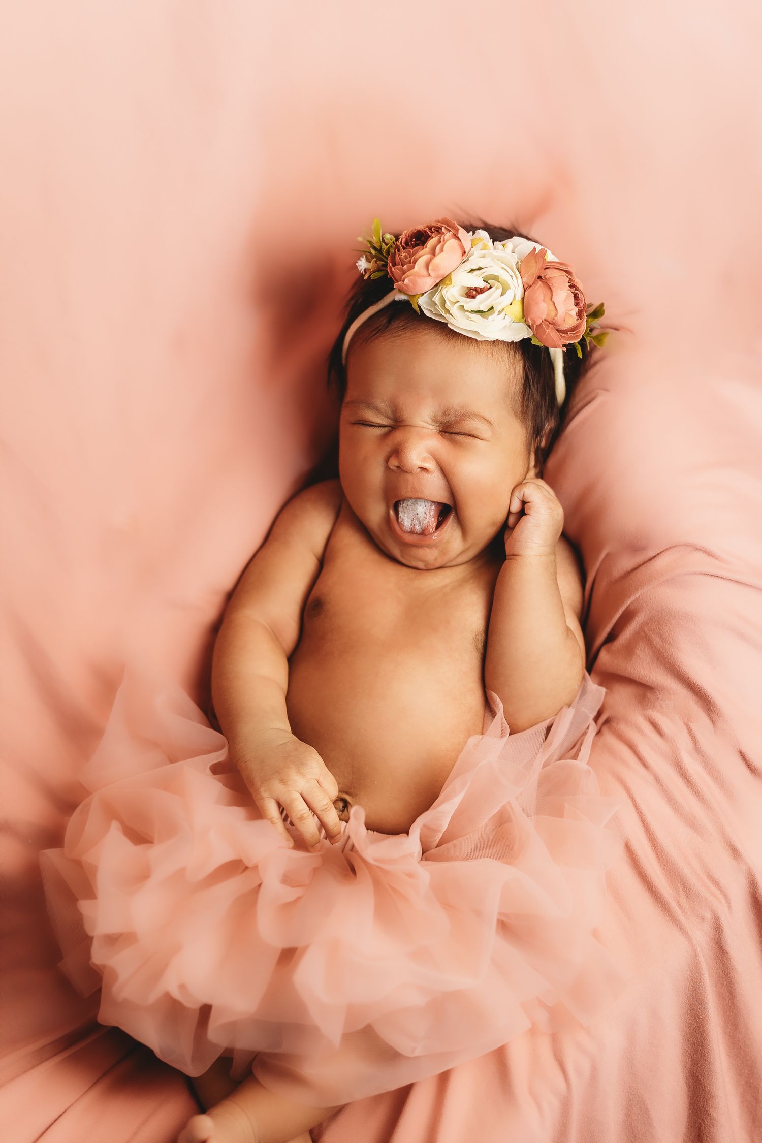 Yawning newborn baby girl in a pink tutu and floral headband resting on a soft pink backdrop.