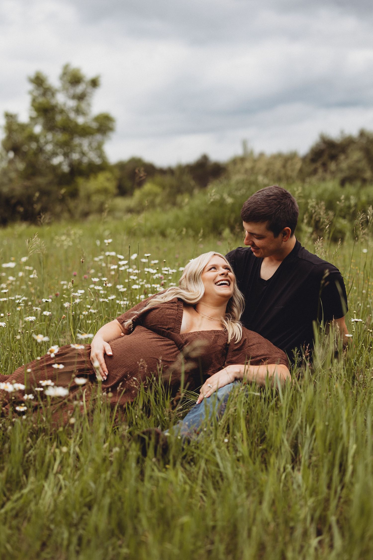 A laughing couple sits together in a wildflower meadow, the woman reclining against the man in black.